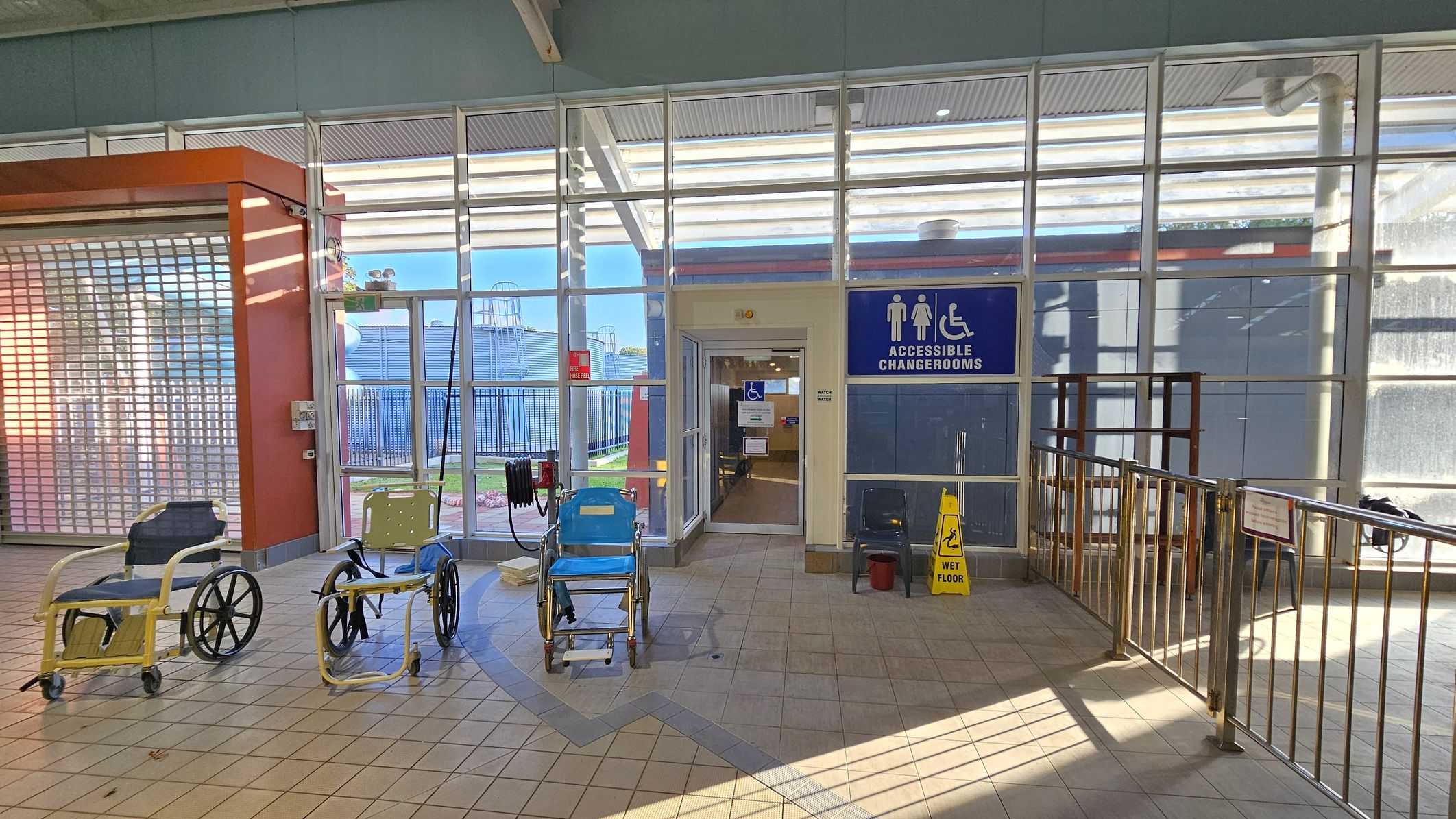 A tiled floor area with three wheelchairs positioned in front of a glass wall. The wall has a door leading to accessible changerooms, indicated by a blue sign with symbols for male, female, and wheelchair access. A yellow "wet floor" sign is placed near the entrance. To the left, there's an orange structure with a metal shutter. Metal railings are on the right side, and outside, a fenced area with some structures is visible through the glass. A tiled floor area with three wheelchairs positioned in front of a glass wall. The wall has a door leading to accessible changerooms, indicated by a blue sign with symbols for male, female, and wheelchair access. A yellow "wet floor" sign is placed near the entrance. To the left, there's an orange structure with a metal shutter. Metal railings are on the right side, and outside, a fenced area with some structures is visible through the glass.