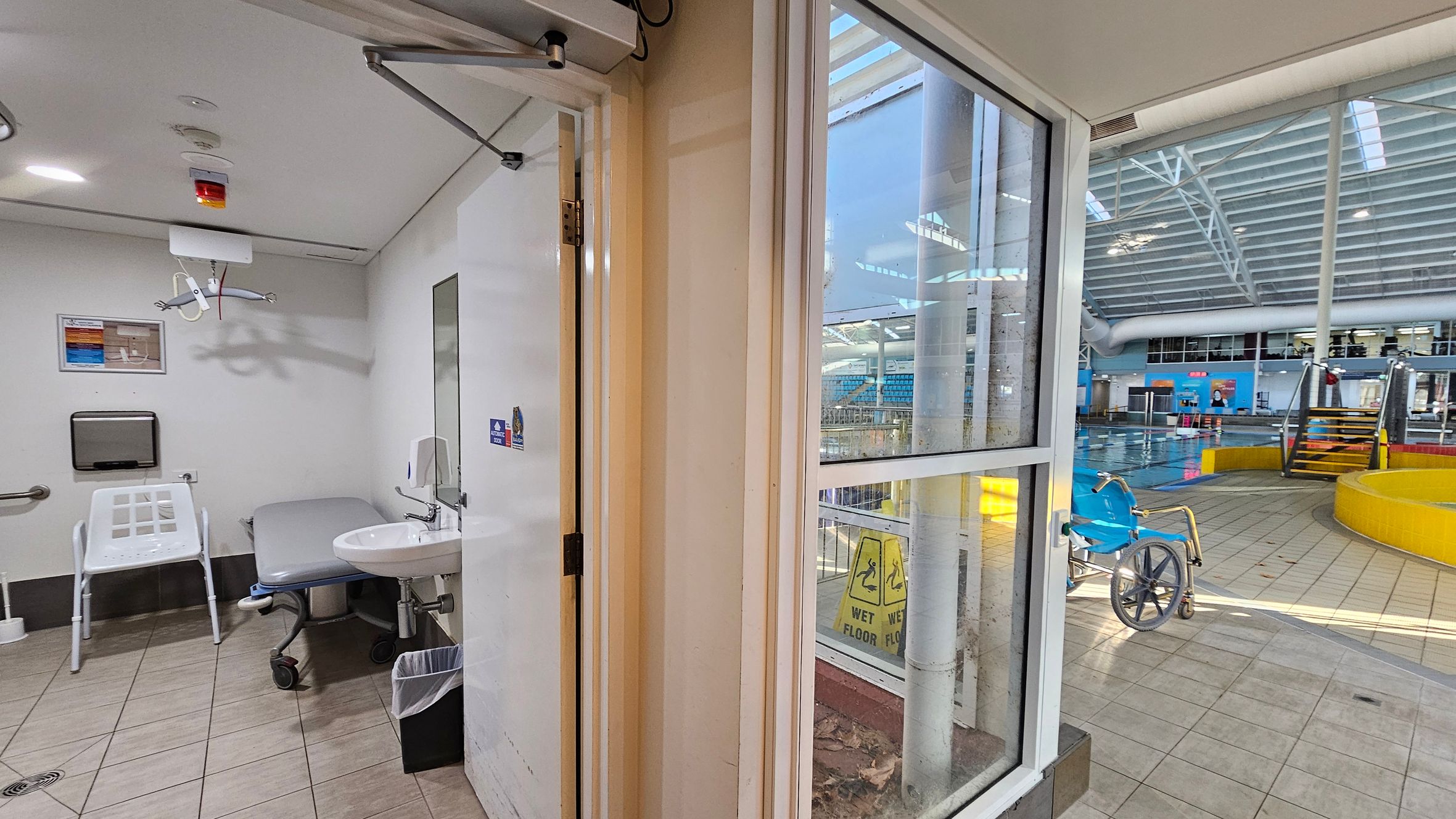 On the left, a changing room with a white chair, a sink, a mirror, and a paper towel dispenser. The ceiling has a hoist system. On the right, a swimming pool area with a blue wheelchair, yellow barriers, and steps leading into the pool. The floor is tiled, and large windows separate the two areas. A "Wet Floor" sign is visible through the window. The ceiling in the pool area has a grid-like structure with skylights. On the left, a changing room with a white chair, a sink, a mirror, and a paper towel dispenser. The ceiling has a hoist system. On the right, a swimming pool area with a blue wheelchair, yellow barriers, and steps leading into the pool. The floor is tiled, and large windows separate the two areas. A "Wet Floor" sign is visible through the window. The ceiling in the pool area has a grid-like structure with skylights.