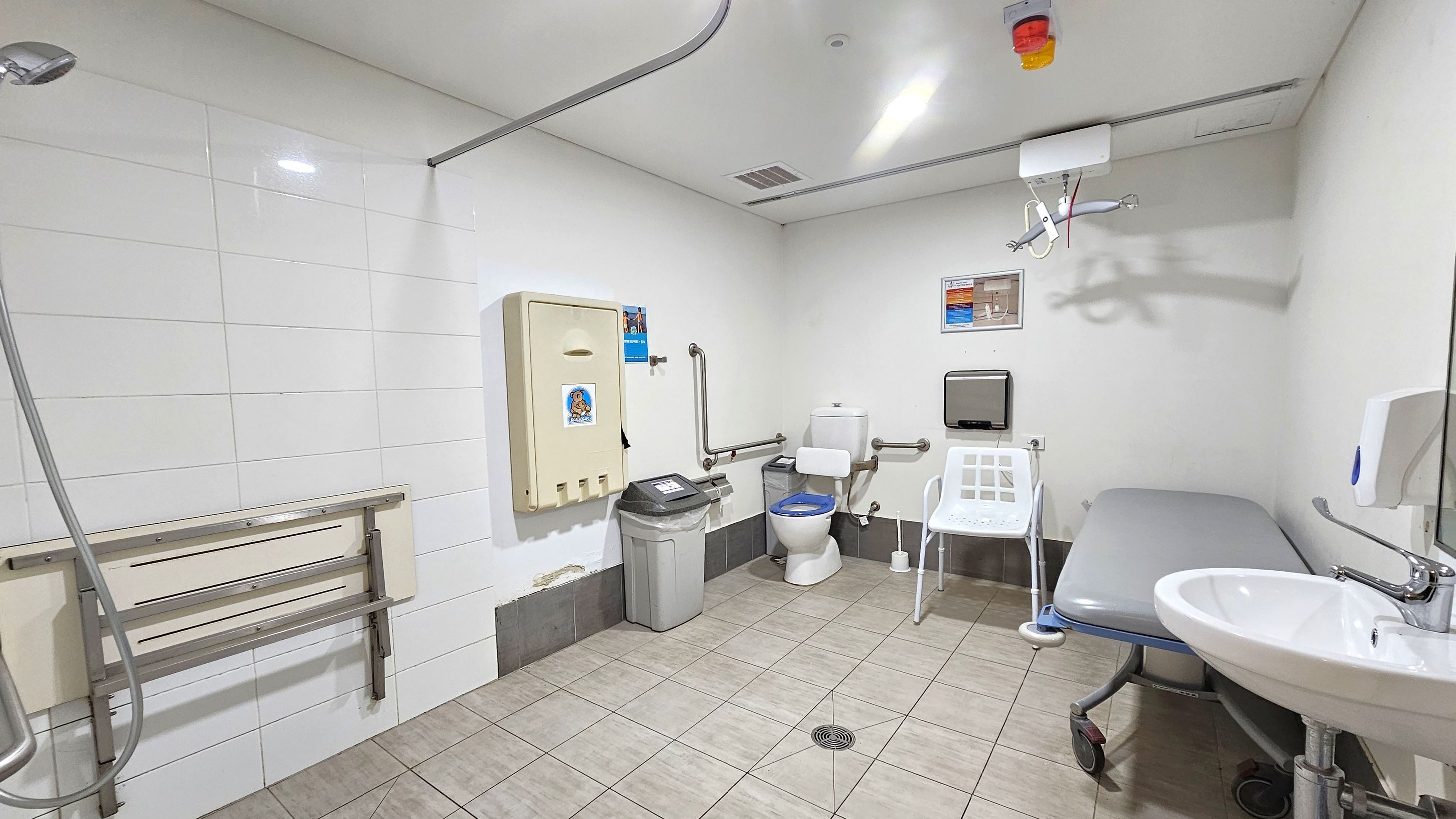 A spacious accessible bathroom with white tiled walls and a light green tiled floor. On the left, a showerhead and a foldable shower seat are mounted on the wall. In the centre, a toilet with support rails, a nappy changing station, and a waste bin are visible. On the right, a white chair, a grey padded changing table on wheels, and a sink with a soap dispenser are present. The ceiling has a hoist system and a light fixture. A spacious accessible bathroom with white tiled walls and a light green tiled floor. On the left, a showerhead and a foldable shower seat are mounted on the wall. In the centre, a toilet with support rails, a nappy changing station, and a waste bin are visible. On the right, a white chair, a grey padded changing table on wheels, and a sink with a soap dispenser are present. The ceiling has a hoist system and a light fixture.