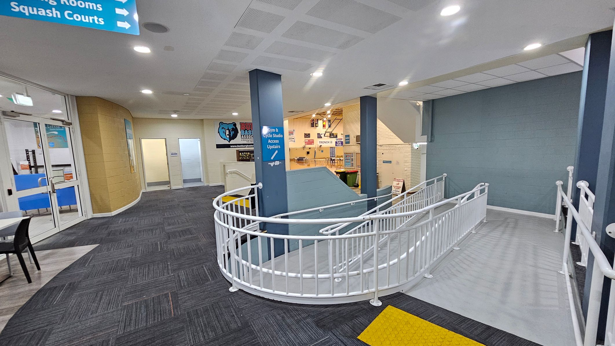 A hallway with a grey carpeted floor and a white ceiling with recessed lights. On the left, there are glass doors leading to a room with blue chairs. Signs above indicate directions to "Squash Courts" and "Change Rooms." In the centre, a blue pillar has a sign for "Gym & Cycle Studio Access Upstairs." A white railing curves around a ramp leading downwards. The walls are a mix of beige and blue, with a logo featuring a bear on the wall. A hallway with a grey carpeted floor and a white ceiling with recessed lights. On the left, there are glass doors leading to a room with blue chairs. Signs above indicate directions to "Squash Courts" and "Change Rooms." In the centre, a blue pillar has a sign for "Gym & Cycle Studio Access Upstairs." A white railing curves around a ramp leading downwards. The walls are a mix of beige and blue, with a logo featuring a bear on the wall.
