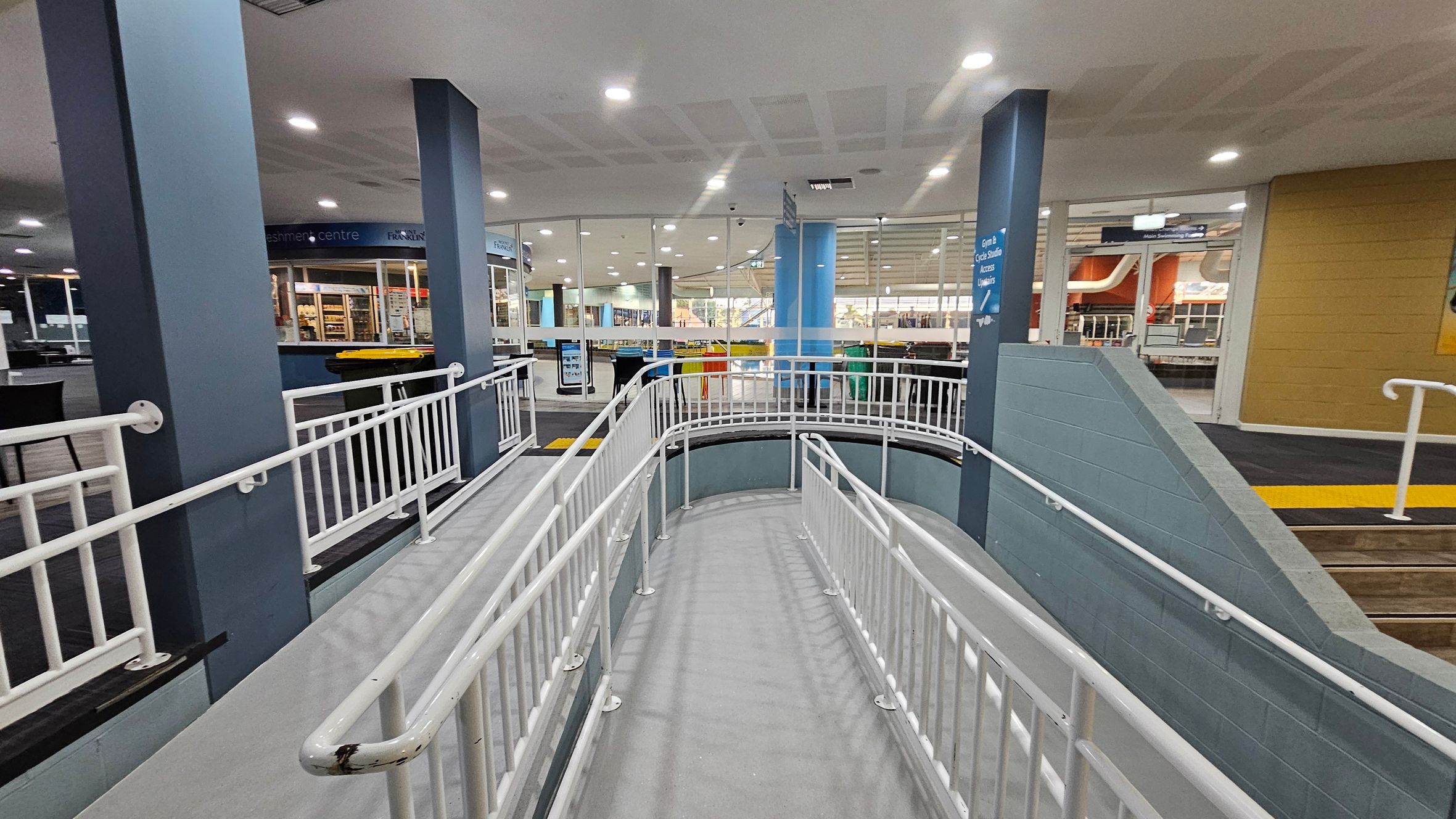 A spacious indoor area with a ramp in the centre, featuring white railings. The floor is grey, and the ceiling is white with recessed lighting. On the left, there are dark blue pillars and a refreshment centre with a counter displaying items. The right side has a yellow wall and a staircase with a yellow tactile strip. In the background, glass walls reveal more of the interior, including seating and additional structures. A spacious indoor area with a ramp in the centre, featuring white railings. The floor is grey, and the ceiling is white with recessed lighting. On the left, there are dark blue pillars and a refreshment centre with a counter displaying items. The right side has a yellow wall and a staircase with a yellow tactile strip. In the background, glass walls reveal more of the interior, including seating and additional structures.