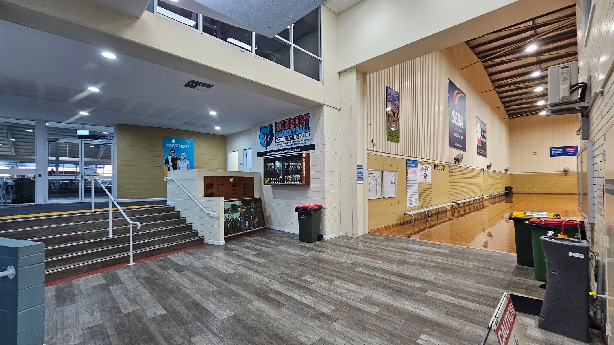 A spacious indoor area with a grey and brown tiled floor. On the left, a staircase with white railings leads up to a glass entrance. A poster with two people is visible on the wall. In the centre, a display case with trophies and a sign for a basketball association. On the right, a wooden-panelled wall with benches and several posters. The ceiling is high with exposed beams and lights. Several bins and a caution sign are in the foreground. A spacious indoor area with a grey and brown tiled floor. On the left, a staircase with white railings leads up to a glass entrance. A poster with two people is visible on the wall. In the centre, a display case with trophies and a sign for a basketball association. On the right, a wooden-panelled wall with benches and several posters. The ceiling is high with exposed beams and lights. Several bins and a caution sign are in the foreground.