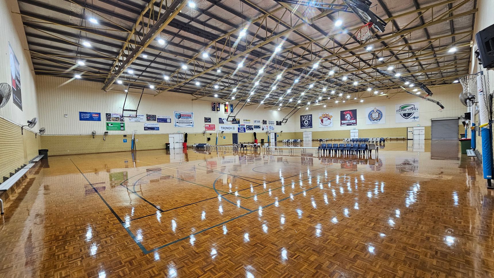 A spacious indoor sports hall with a polished wooden floor marked for basketball. On the left, there are benches and wall-mounted fans. The right side features a row of stacked chairs and a basketball hoop. The back wall displays various banners and logos. The ceiling is high with exposed beams and numerous bright lights. A spacious indoor sports hall with a polished wooden floor marked for basketball. On the left, there are benches and wall-mounted fans. The right side features a row of stacked chairs and a basketball hoop. The back wall displays various banners and logos. The ceiling is high with exposed beams and numerous bright lights.