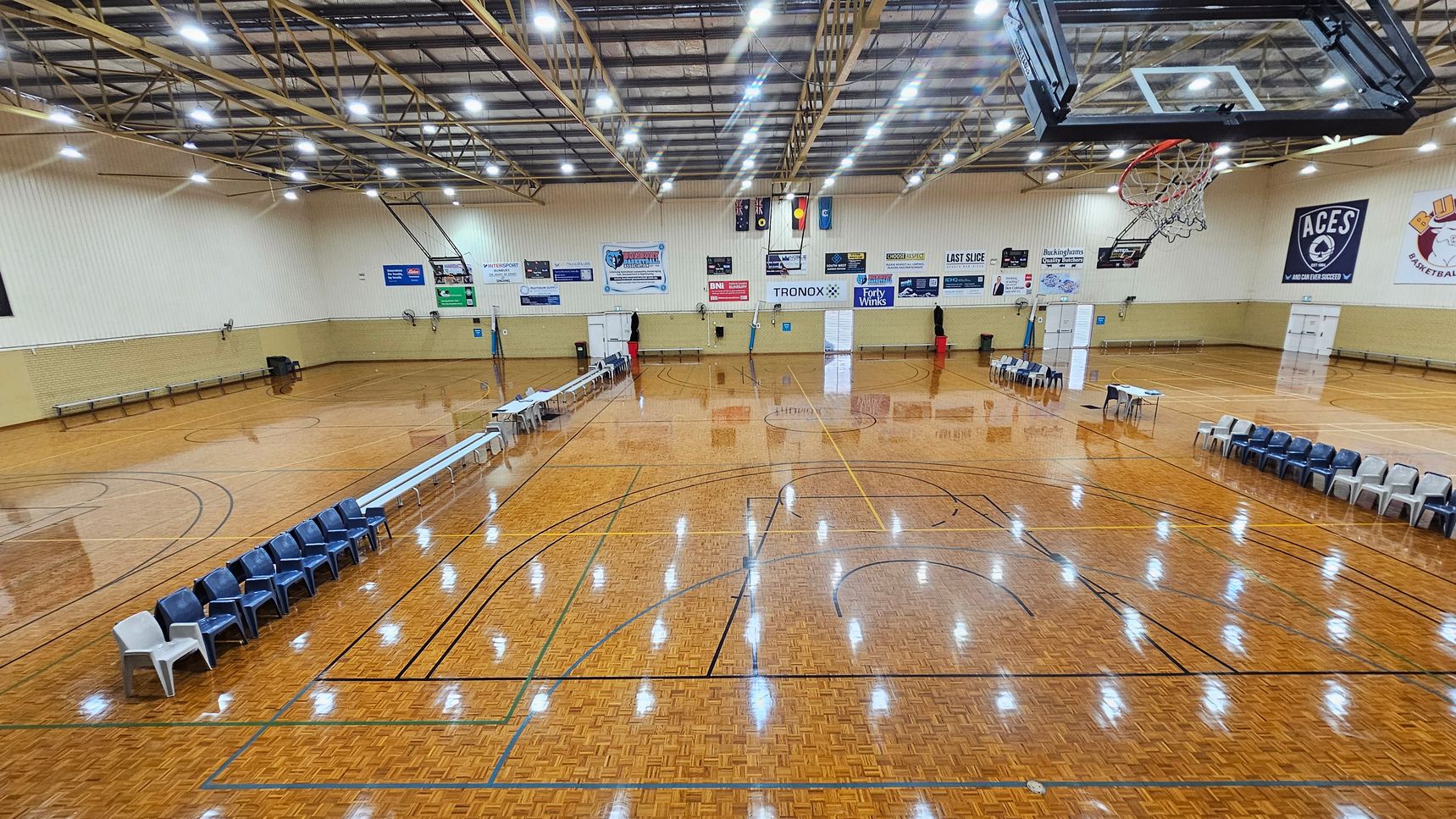 A large indoor basketball court with a polished wooden floor. On the left, there are rows of blue and white chairs along the sideline. In the centre, the court markings are visible, and a scoreboard is mounted on the far wall. On the right, more chairs are arranged, and a basketball hoop is suspended from the ceiling. The ceiling is high with exposed beams and bright overhead lights. Various banners and advertisements are displayed on the walls. A large indoor basketball court with a polished wooden floor. On the left, there are rows of blue and white chairs along the sideline. In the centre, the court markings are visible, and a scoreboard is mounted on the far wall. On the right, more chairs are arranged, and a basketball hoop is suspended from the ceiling. The ceiling is high with exposed beams and bright overhead lights. Various banners and advertisements are displayed on the walls.