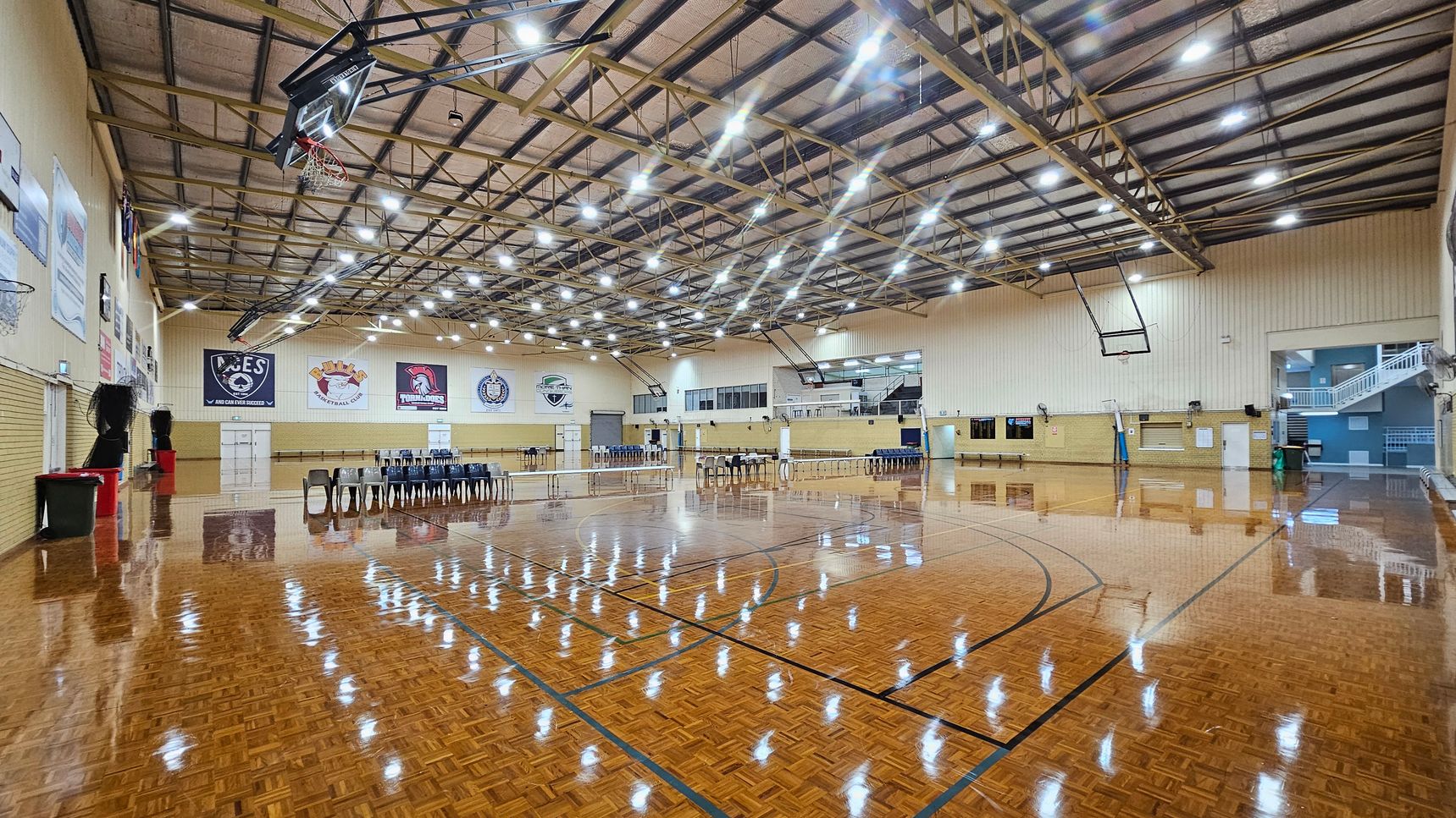 A spacious indoor sports hall with a polished wooden floor marked for basketball. On the left, there are basketball hoops and banners on the walls. In the centre, rows of black chairs are arranged, and a volleyball net is set up. The right side features more chairs and a staircase leading to an upper level. The ceiling is high with exposed beams and numerous bright lights. The walls are light-coloured, and there are additional banners and doors visible. A spacious indoor sports hall with a polished wooden floor marked for basketball. On the left, there are basketball hoops and banners on the walls. In the centre, rows of black chairs are arranged, and a volleyball net is set up. The right side features more chairs and a staircase leading to an upper level. The ceiling is high with exposed beams and numerous bright lights. The walls are light-coloured, and there are additional banners and doors visible.