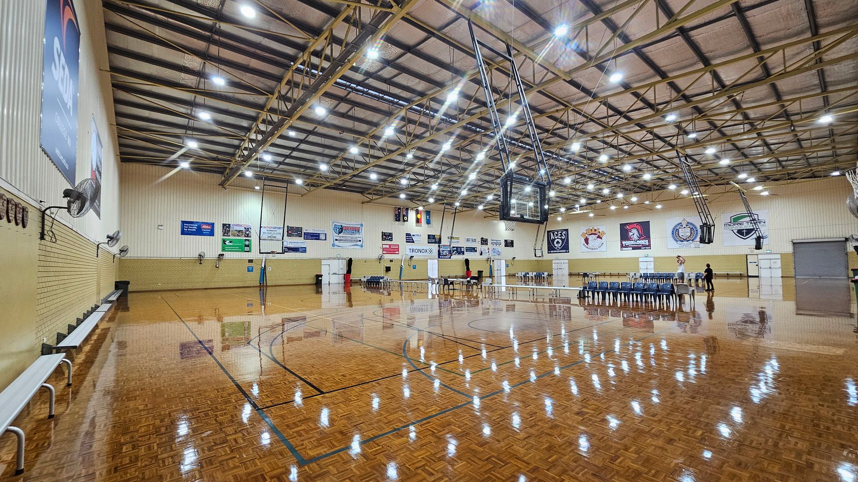 A large indoor sports hall with a polished wooden floor reflecting the overhead lights. On the left, there are benches against a brick wall with several large fans mounted above. The ceiling is high with exposed beams and numerous lights. In the centre, a basketball hoop is suspended. On the right, there are rows of stacked chairs and a few people standing near tables. The walls are adorned with various banners and logos. A large indoor sports hall with a polished wooden floor reflecting the overhead lights. On the left, there are benches against a brick wall with several large fans mounted above. The ceiling is high with exposed beams and numerous lights. In the centre, a basketball hoop is suspended. On the right, there are rows of stacked chairs and a few people standing near tables. The walls are adorned with various banners and logos.