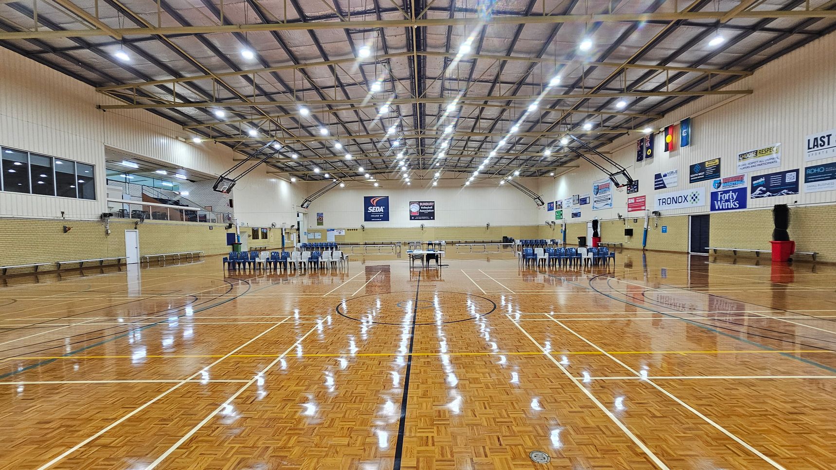 A large indoor sports hall with a polished wooden floor marked for various sports. In the centre, there are rows of blue chairs and a table. The walls are lined with banners and advertisements. The ceiling is high with exposed beams and bright overhead lights. On the left, there are windows and a small upper viewing area. On the right, more chairs are arranged, and there are flags displayed on the wall. The space is open and well-lit. A large indoor sports hall with a polished wooden floor marked for various sports. In the centre, there are rows of blue chairs and a table. The walls are lined with banners and advertisements. The ceiling is high with exposed beams and bright overhead lights. On the left, there are windows and a small upper viewing area. On the right, more chairs are arranged, and there are flags displayed on the wall. The space is open and well-lit.