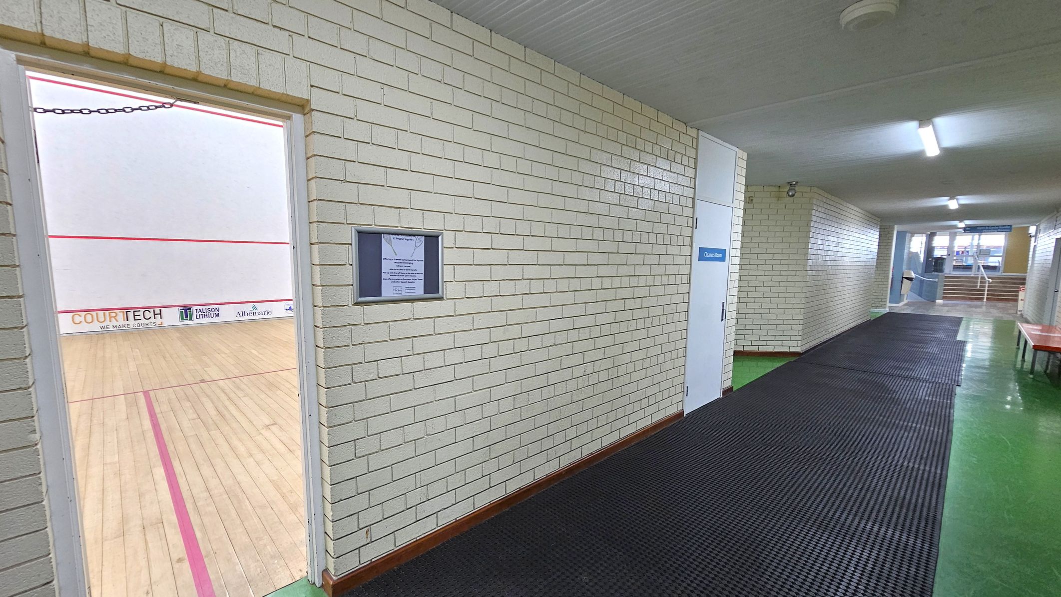 A corridor with white brick walls and a green floor. On the left, an open door reveals a squash court with a wooden floor and red boundary lines. The court has advertisements on the lower wall. On the right, a white door labelled "Cleaner Room" is visible. The corridor has a black rubber mat running along the floor. In the background, there are stairs leading up, with windows allowing natural light to enter. The ceiling is white with fluorescent lights. A corridor with white brick walls and a green floor. On the left, an open door reveals a squash court with a wooden floor and red boundary lines. The court has advertisements on the lower wall. On the right, a white door labelled "Cleaner Room" is visible. The corridor has a black rubber mat running along the floor. In the background, there are stairs leading up, with windows allowing natural light to enter. The ceiling is white with fluorescent lights.