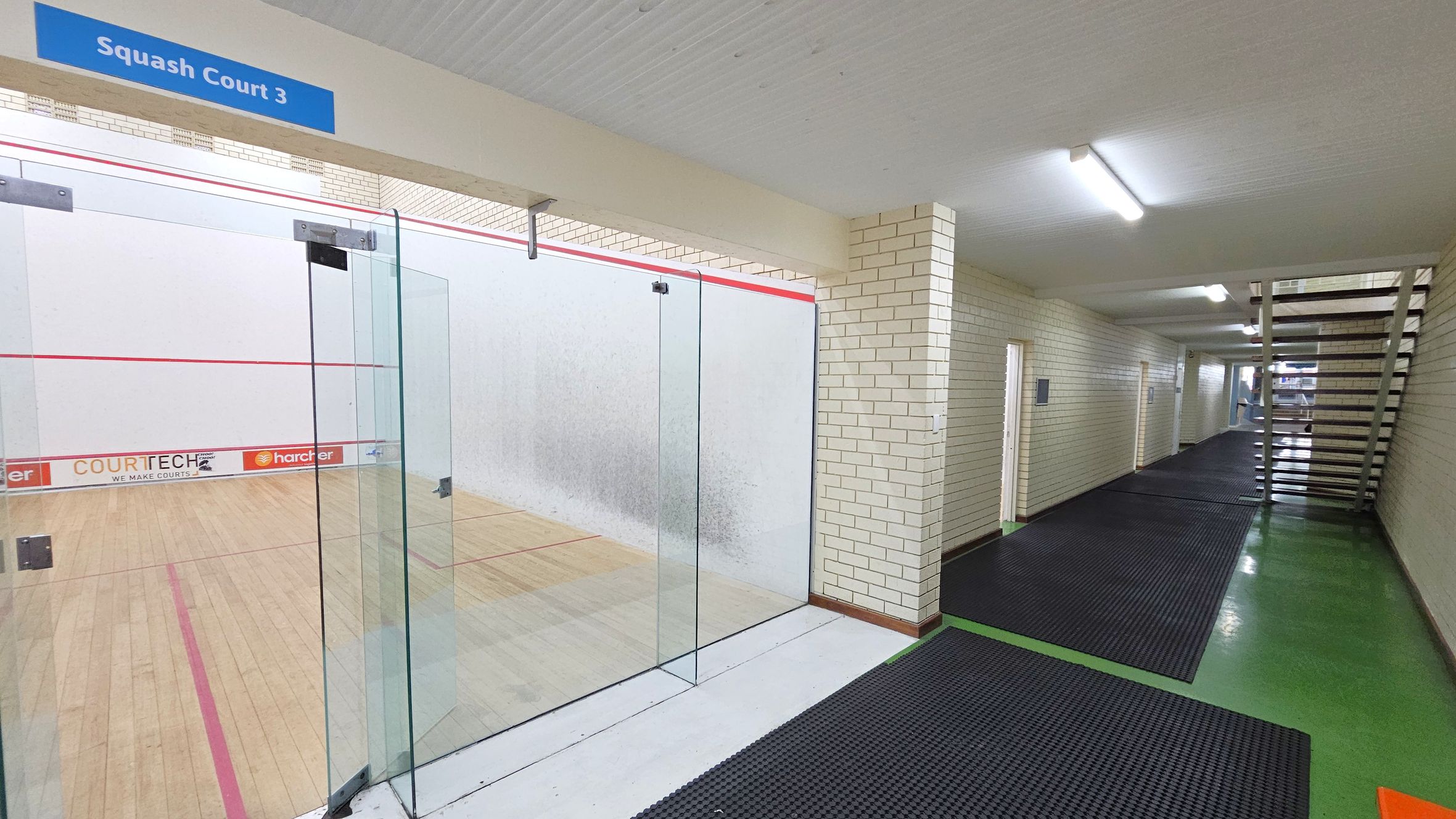 A squash court is on the left, enclosed by glass walls with a sign above reading "Squash Court 3". The court has a wooden floor with red boundary lines. On the right, a corridor with cream brick walls and black rubber mats on a green floor leads to a staircase. The ceiling is white with fluorescent lights. A squash court is on the left, enclosed by glass walls with a sign above reading "Squash Court 3". The court has a wooden floor with red boundary lines. On the right, a corridor with cream brick walls and black rubber mats on a green floor leads to a staircase. The ceiling is white with fluorescent lights.