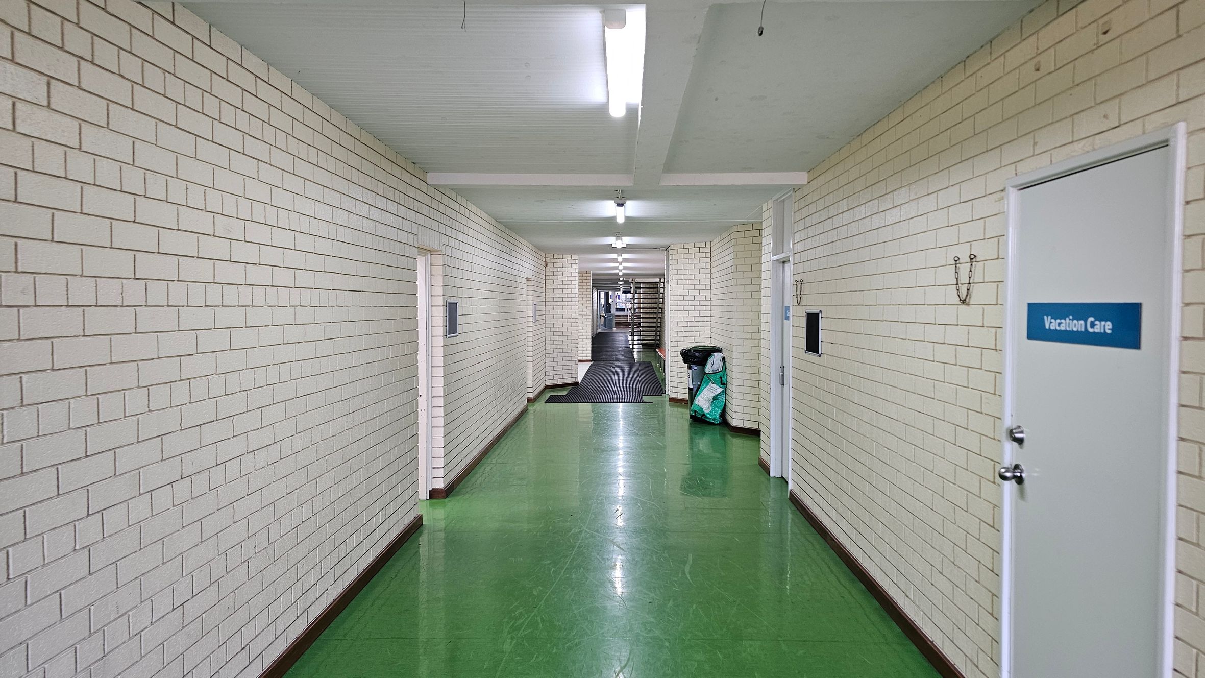 A long corridor with white brick walls and a shiny green floor. On the right, there is a door labelled "Vacation Care" and a black bin with a bag beside it. The ceiling is white with fluorescent lights evenly spaced along the length of the corridor. In the distance, the corridor narrows and leads to a staircase. The walls have hooks and small black panels mounted on them. A long corridor with white brick walls and a shiny green floor. On the right, there is a door labelled "Vacation Care" and a black bin with a bag beside it. The ceiling is white with fluorescent lights evenly spaced along the length of the corridor. In the distance, the corridor narrows and leads to a staircase. The walls have hooks and small black panels mounted on them.