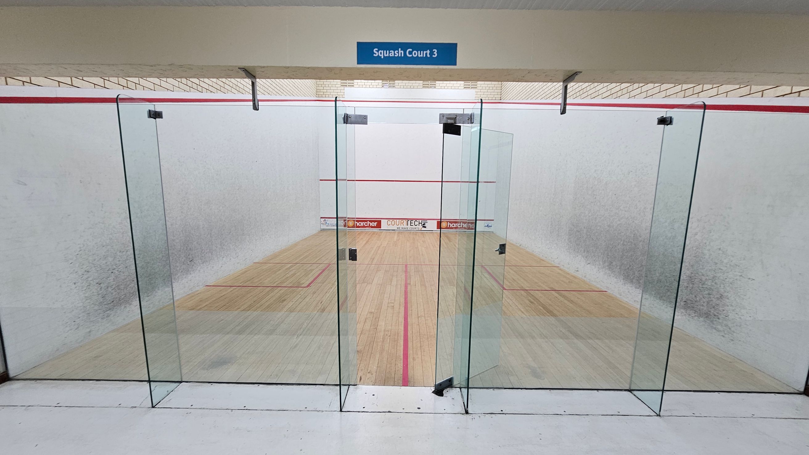 A squash court with a glass door entrance labelled "Squash Court 3" at the top. The court has wooden flooring with red boundary lines. The walls are white with a red line running horizontally near the top. Advertisements are visible at the far end of the court. The ceiling is light-coloured with visible beams. A squash court with a glass door entrance labelled "Squash Court 3" at the top. The court has wooden flooring with red boundary lines. The walls are white with a red line running horizontally near the top. Advertisements are visible at the far end of the court. The ceiling is light-coloured with visible beams.