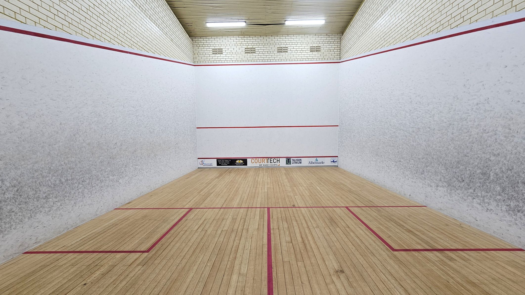 A squash court with light wooden flooring marked by red lines. The walls are white with a red boundary line near the top. The back wall features several sponsor logos at the bottom. The ceiling is wooden with fluorescent lights. The side walls have a slightly textured appearance. A squash court with light wooden flooring marked by red lines. The walls are white with a red boundary line near the top. The back wall features several sponsor logos at the bottom. The ceiling is wooden with fluorescent lights. The side walls have a slightly textured appearance.