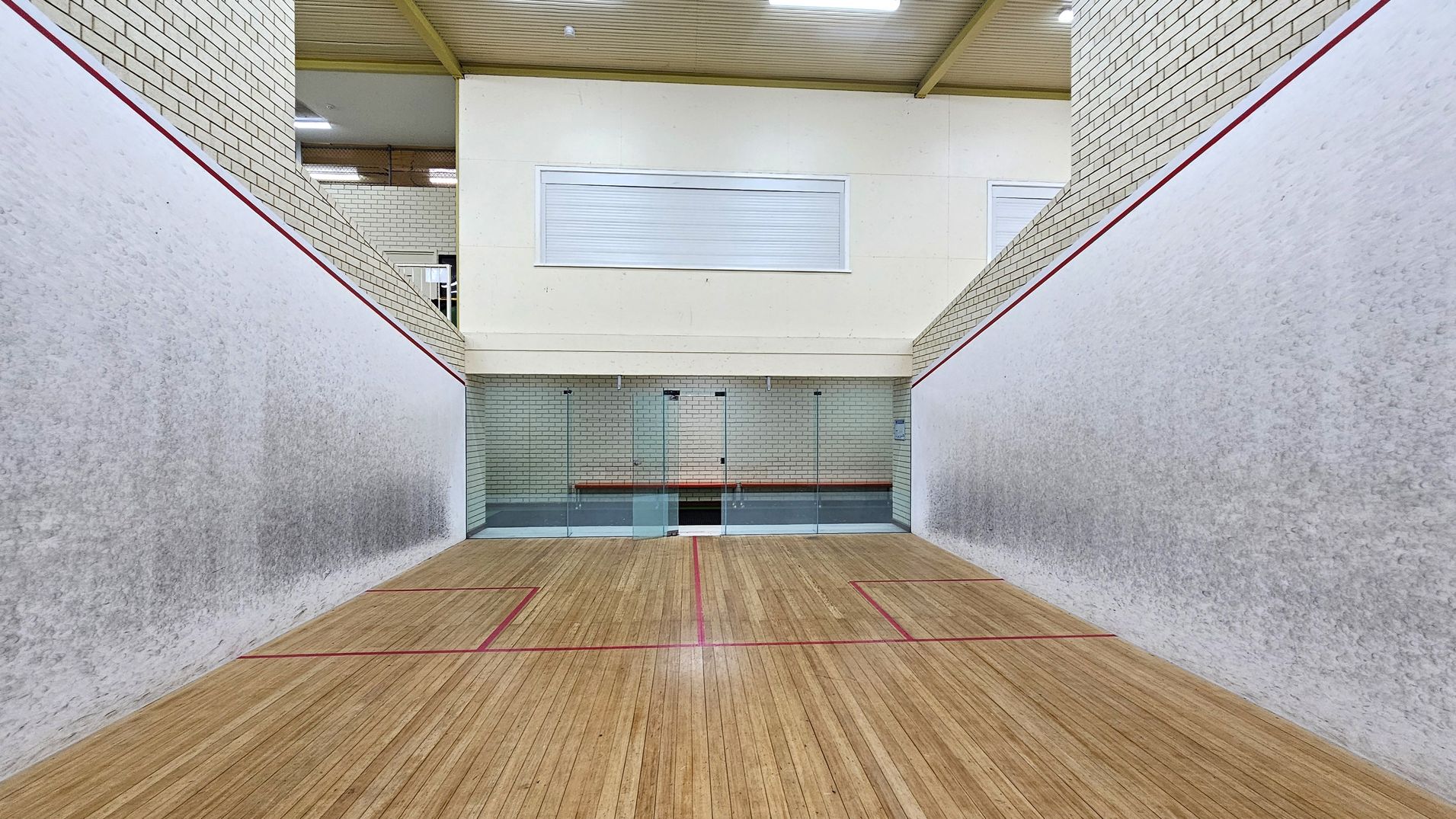 A squash court with wooden flooring marked by red lines. The walls are light grey with a red outline near the top. The back wall is made of glass panels, allowing a view into an adjacent area with white brick walls. Above the glass, there is a white panel with a closed window. The ceiling is wooden with visible beams and fluorescent lights. A squash court with wooden flooring marked by red lines. The walls are light grey with a red outline near the top. The back wall is made of glass panels, allowing a view into an adjacent area with white brick walls. Above the glass, there is a white panel with a closed window. The ceiling is wooden with visible beams and fluorescent lights.