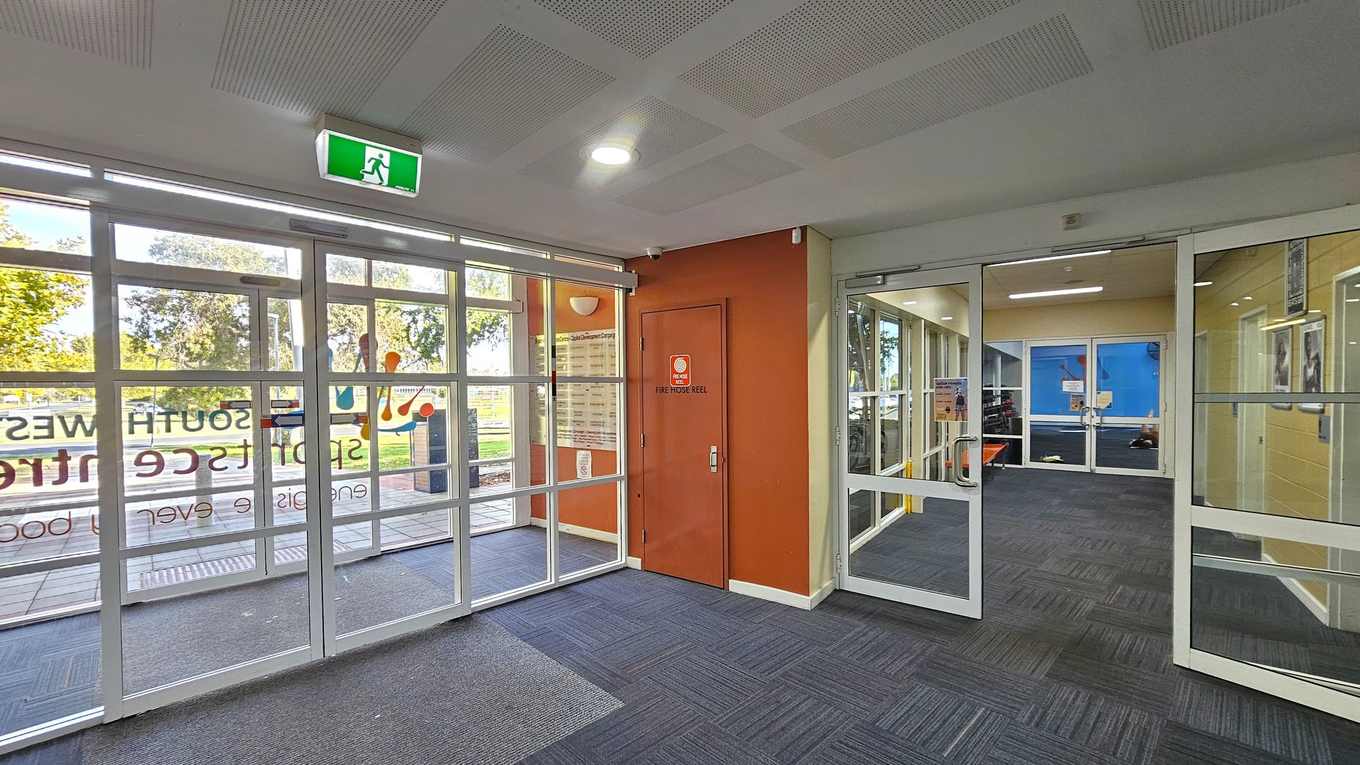 A modern interior space with glass doors on the left, featuring signage and a view of trees outside. In the centre, an orange wall with a door marked "Fire Hose Reel". On the right, more glass doors lead to another room with blue walls. The floor is covered with dark grey and blue patterned carpet tiles, and the ceiling has a grid pattern with recessed lighting. An illuminated green exit sign is above the left doors. A modern interior space with glass doors on the left, featuring signage and a view of trees outside. In the centre, an orange wall with a door marked "Fire Hose Reel". On the right, more glass doors lead to another room with blue walls. The floor is covered with dark grey and blue patterned carpet tiles, and the ceiling has a grid pattern with recessed lighting. An illuminated green exit sign is above the left doors.