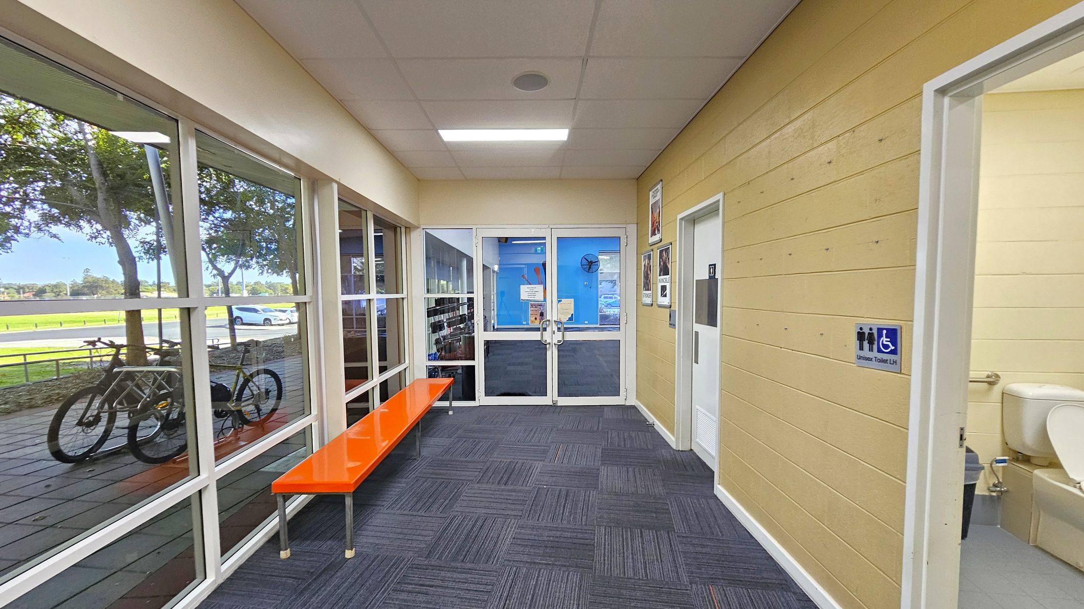 A hallway with a glass wall on the left, showing a view of trees, a parked bicycle, and a car outside. An orange bench is positioned along the glass wall. The right side has a light yellow wooden wall with a door leading to a unisex toilet, partially visible. The floor is covered with blue patterned carpet tiles. At the end of the hallway, there are double glass doors with notices and a blue wall visible through them. The ceiling is fitted with a fluorescent light. A hallway with a glass wall on the left, showing a view of trees, a parked bicycle, and a car outside. An orange bench is positioned along the glass wall. The right side has a light yellow wooden wall with a door leading to a unisex toilet, partially visible. The floor is covered with blue patterned carpet tiles. At the end of the hallway, there are double glass doors with notices and a blue wall visible through them. The ceiling is fitted with a fluorescent light.