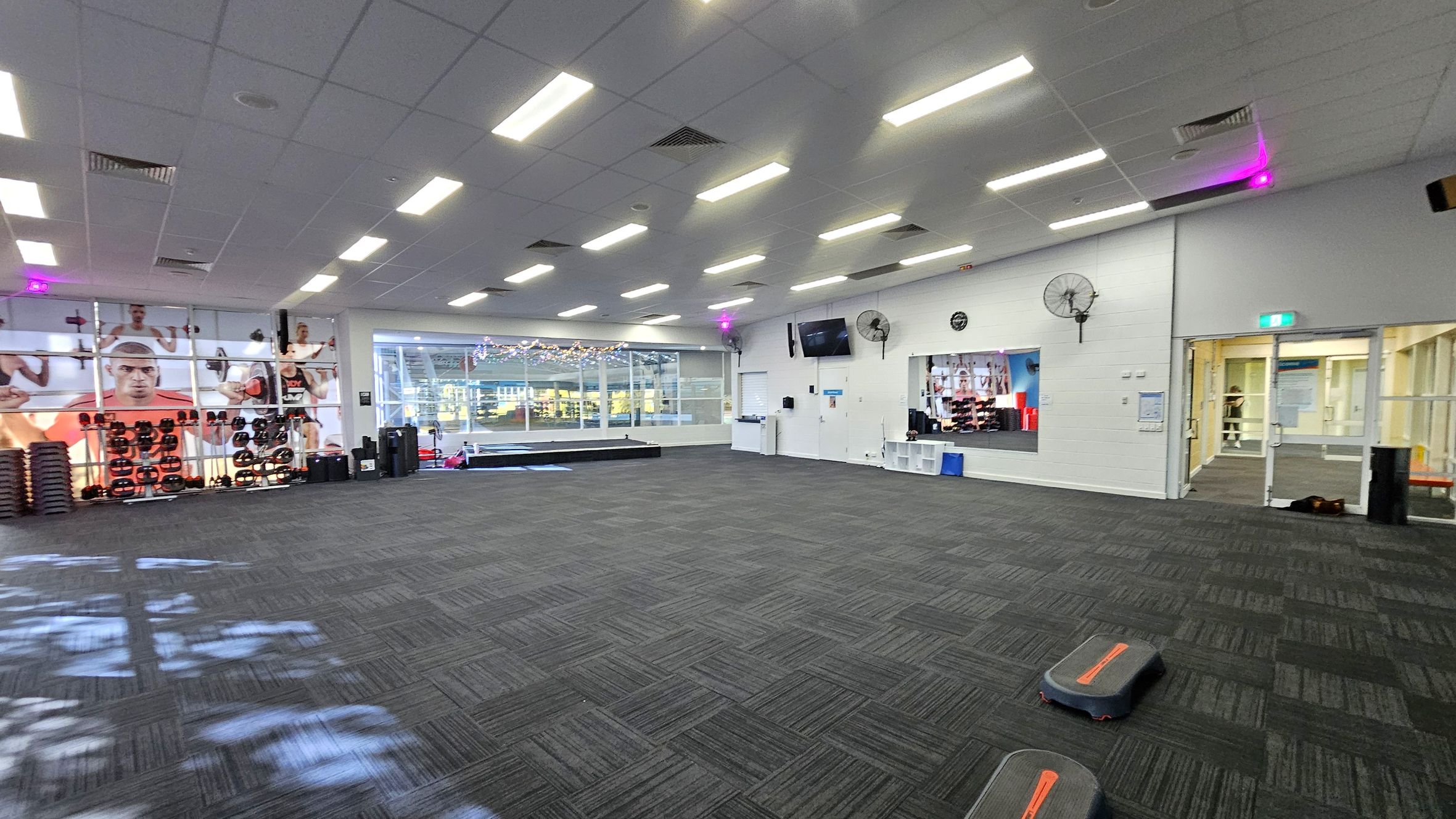 A spacious gym room with a dark carpeted floor. On the left, there are shelves with weights and a large wall mural of people exercising. The back wall features large windows with a view of an outdoor area, decorated with string lights. On the right, a wall-mounted TV, fans, and a mirror are visible. There are exercise steps on the floor. The ceiling is lined with fluorescent lights. A glass door on the right leads to another area. A spacious gym room with a dark carpeted floor. On the left, there are shelves with weights and a large wall mural of people exercising. The back wall features large windows with a view of an outdoor area, decorated with string lights. On the right, a wall-mounted TV, fans, and a mirror are visible. There are exercise steps on the floor. The ceiling is lined with fluorescent lights. A glass door on the right leads to another area.