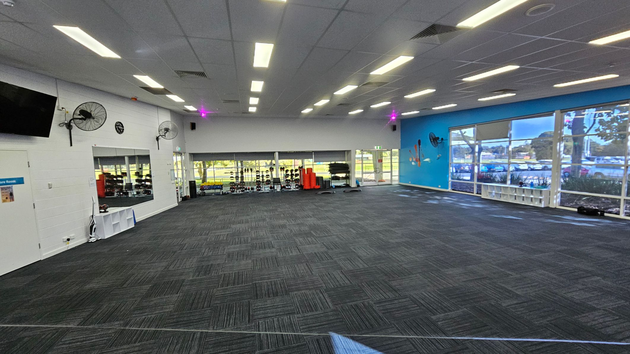 A spacious gym room with a dark carpeted floor and a white ceiling with fluorescent lights. On the left, there's a wall with a mounted TV, two fans, and a clock. Below, a door and a window reveal gym equipment inside. The back wall has large windows showing outdoor greenery and more gym equipment like weights and mats. The right wall is blue with large windows, allowing natural light in. A small shelf is visible beneath the windows. A spacious gym room with a dark carpeted floor and a white ceiling with fluorescent lights. On the left, there's a wall with a mounted TV, two fans, and a clock. Below, a door and a window reveal gym equipment inside. The back wall has large windows showing outdoor greenery and more gym equipment like weights and mats. The right wall is blue with large windows, allowing natural light in. A small shelf is visible beneath the windows.