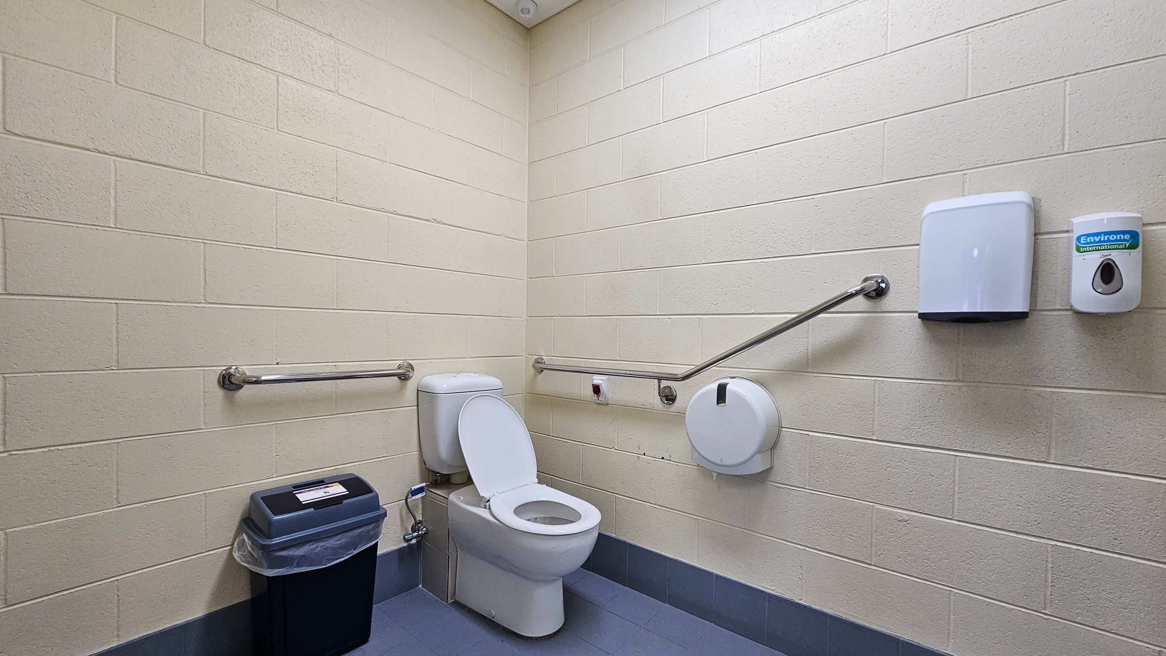 A public restroom with light-coloured brick walls and a blue-tiled floor. In the centre, a white toilet with the seat up is positioned against the corner. To the left, a black bin with a liner is placed on the floor. On the right wall, a toilet paper dispenser, a paper towel dispenser, and a soap dispenser are mounted. Metal grab bars are installed on both walls for accessibility. The ceiling is plain and light-coloured. A public restroom with light-coloured brick walls and a blue-tiled floor. In the centre, a white toilet with the seat up is positioned against the corner. To the left, a black bin with a liner is placed on the floor. On the right wall, a toilet paper dispenser, a paper towel dispenser, and a soap dispenser are mounted. Metal grab bars are installed on both walls for accessibility. The ceiling is plain and light-coloured.