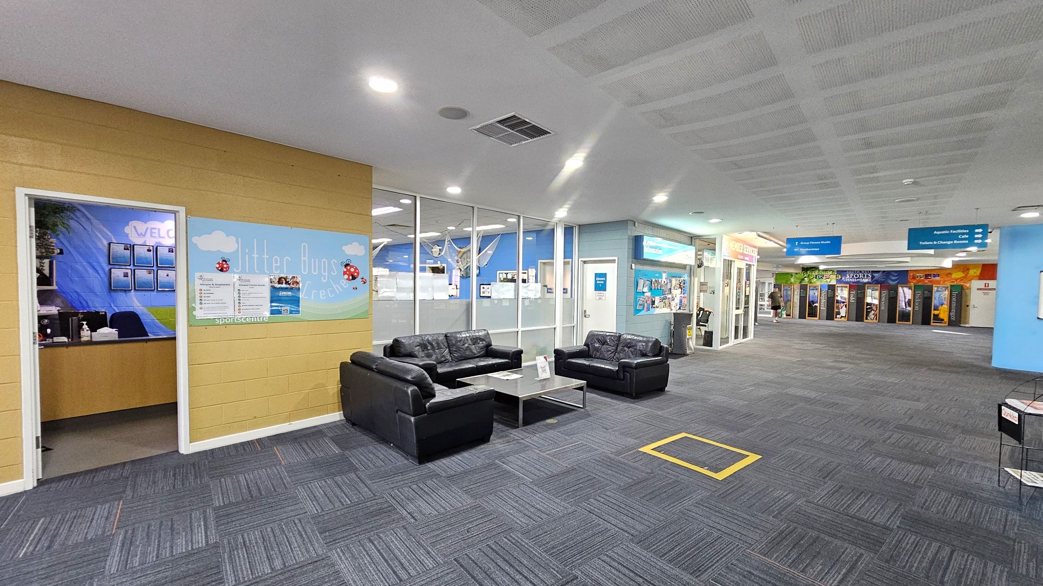 A spacious indoor area with a grey carpeted floor and a white ceiling. On the left, there's a yellow wall with a door leading to an office, featuring a sign with ladybirds. In the centre, a seating area with black leather sofas and a coffee table. The right side has glass walls and doors leading to various facilities, with signs for aquatic facilities and a café. The background shows a corridor with colourful posters and a person walking. A spacious indoor area with a grey carpeted floor and a white ceiling. On the left, there's a yellow wall with a door leading to an office, featuring a sign with ladybirds. In the centre, a seating area with black leather sofas and a coffee table. The right side has glass walls and doors leading to various facilities, with signs for aquatic facilities and a café. The background shows a corridor with colourful posters and a person walking.