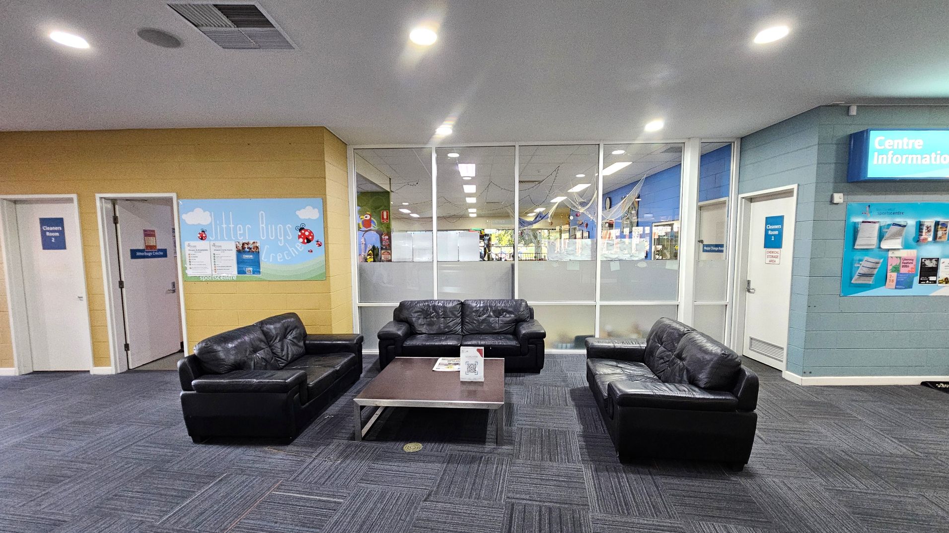 A waiting area with three black leather sofas arranged around a rectangular coffee table in the centre. The floor is covered with dark grey carpet tiles. The back wall features large glass panels, providing a view into another room. On the left, there are two doors labelled "Cleaners Room 2" and "Jitterbugs Crèche" with a colourful sign. On the right, a wall-mounted display titled "Centre Information" is visible. The ceiling is white with recessed lighting. A waiting area with three black leather sofas arranged around a rectangular coffee table in the centre. The floor is covered with dark grey carpet tiles. The back wall features large glass panels, providing a view into another room. On the left, there are two doors labelled "Cleaners Room 2" and "Jitterbugs Crèche" with a colourful sign. On the right, a wall-mounted display titled "Centre Information" is visible. The ceiling is white with recessed lighting.