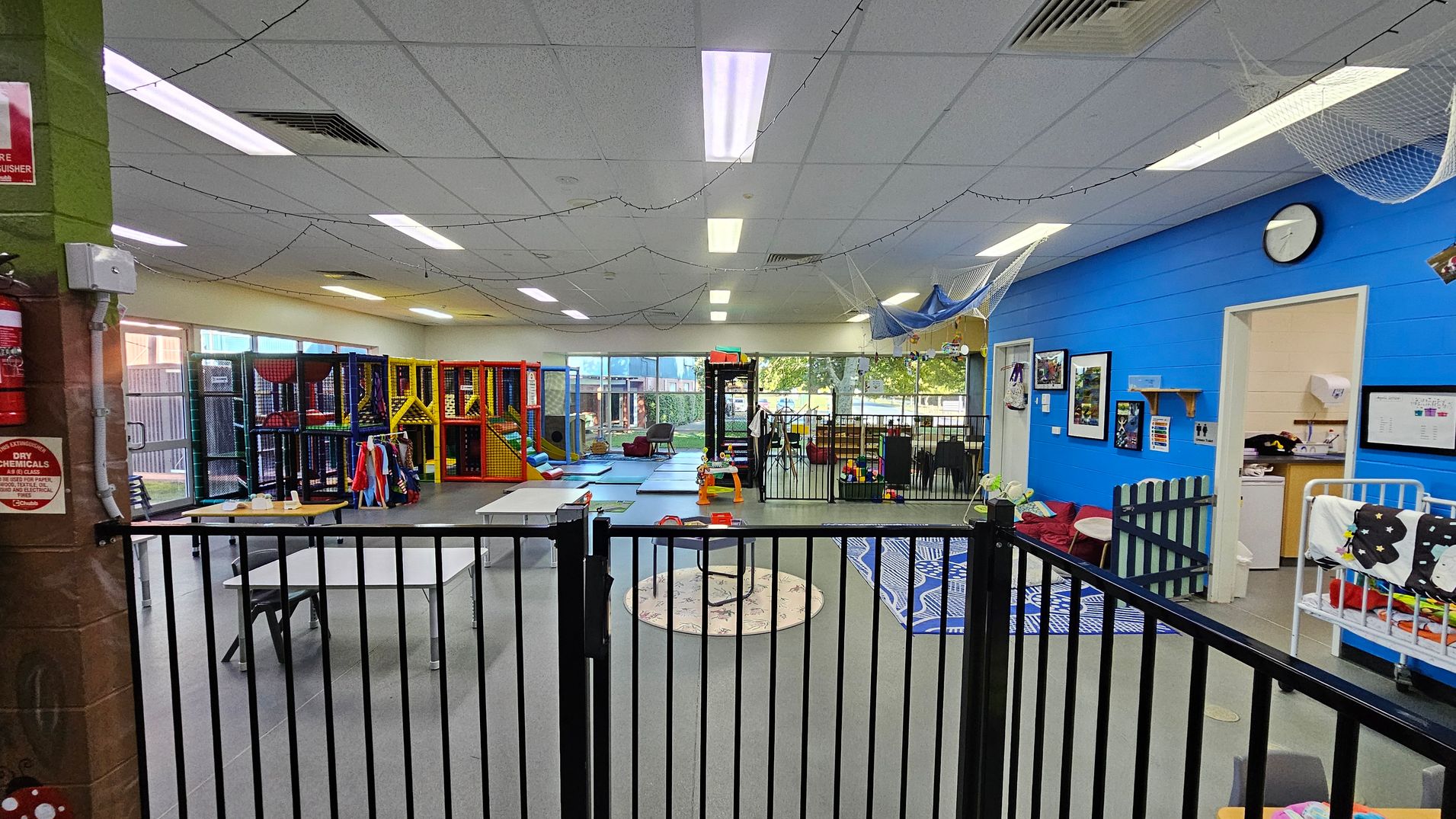 A spacious indoor play area with a grey floor and a ceiling with fluorescent lights. On the left, there are colourful play structures and a table with chairs. In the centre, a circular rug and a small play area are visible. On the right, a blue wall features framed pictures and a clock, with an open door leading to a small room. A black safety gate runs across the foreground, and string lights are draped across the ceiling. A spacious indoor play area with a grey floor and a ceiling with fluorescent lights. On the left, there are colourful play structures and a table with chairs. In the centre, a circular rug and a small play area are visible. On the right, a blue wall features framed pictures and a clock, with an open door leading to a small room. A black safety gate runs across the foreground, and string lights are draped across the ceiling.