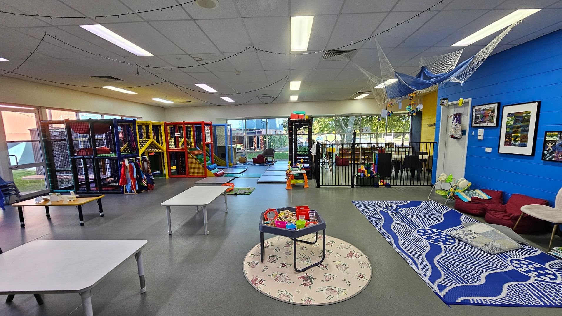 A spacious indoor play area with a grey floor and white ceiling. On the left, there are colourful play structures with slides and climbing areas. In the centre, a small round table with toys sits on a patterned rug. On the right, a blue wall features framed pictures, and a seating area with cushions and a patterned mat. The background has large windows with a view of trees outside, and a fenced-off section with more toys and seating. String lights hang from the ceiling. A spacious indoor play area with a grey floor and white ceiling. On the left, there are colourful play structures with slides and climbing areas. In the centre, a small round table with toys sits on a patterned rug. On the right, a blue wall features framed pictures, and a seating area with cushions and a patterned mat. The background has large windows with a view of trees outside, and a fenced-off section with more toys and seating. String lights hang from the ceiling.