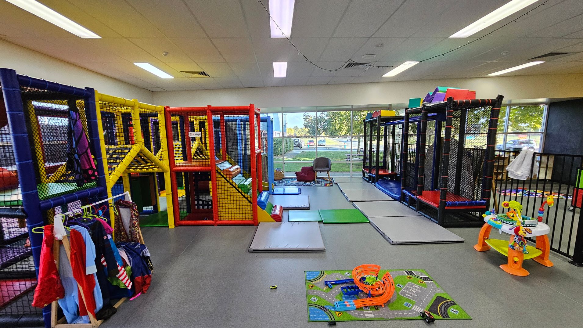Indoor play area with colourful soft play structures on the left, featuring yellow, red, and blue frames with netting. A rack of children's costumes is nearby. In the centre, padded mats are spread on the floor. On the right, a black-framed play structure and a small activity table with toys are visible. Large windows in the background show an outdoor view with trees and parked cars. The ceiling has fluorescent lights and a grey floor. Indoor play area with colourful soft play structures on the left, featuring yellow, red, and blue frames with netting. A rack of children's costumes is nearby. In the centre, padded mats are spread on the floor. On the right, a black-framed play structure and a small activity table with toys are visible. Large windows in the background show an outdoor view with trees and parked cars. The ceiling has fluorescent lights and a grey floor.