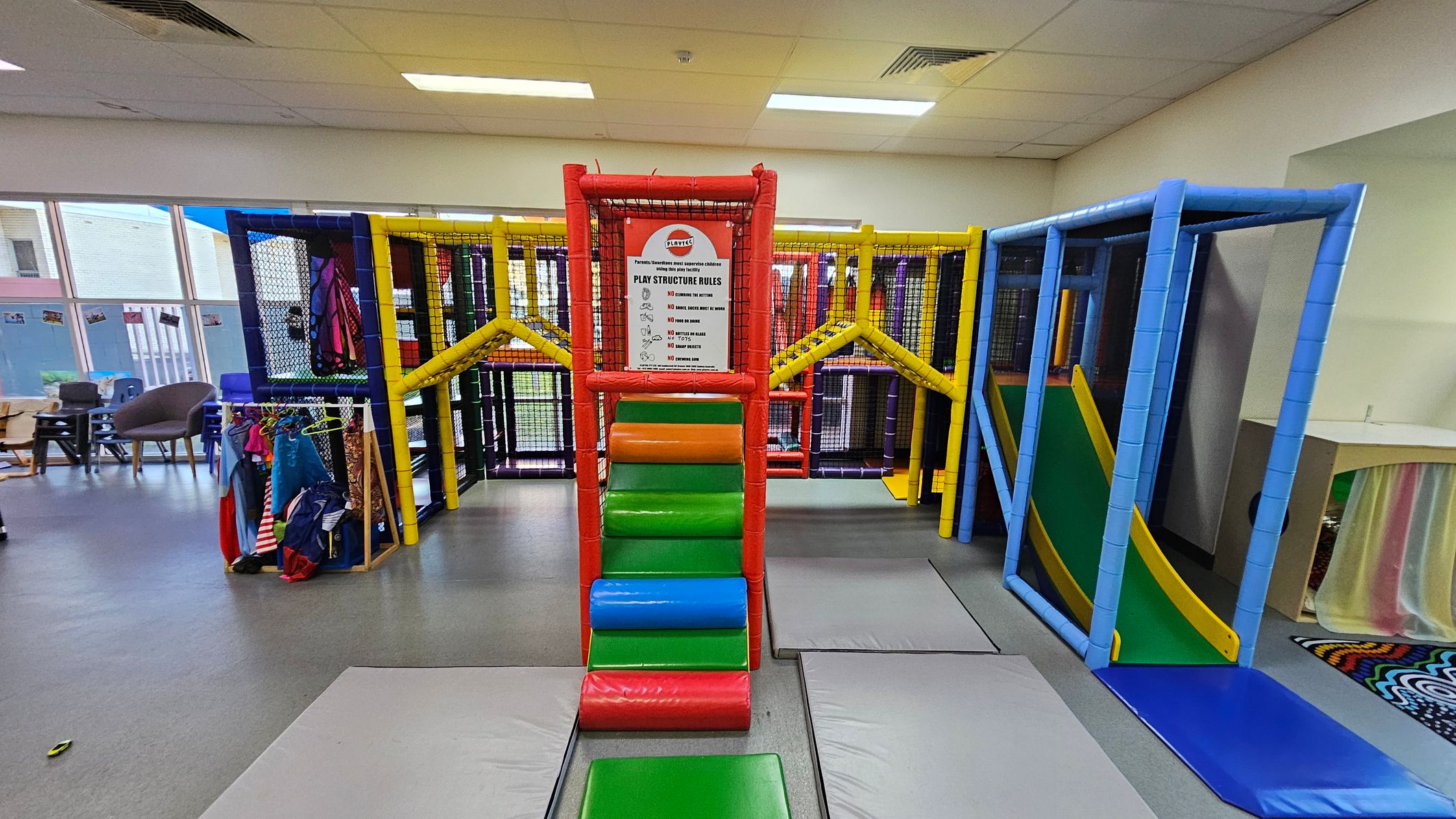 Indoor play area with a colourful climbing structure in the centre, featuring red, green, orange, and blue padded steps leading up to a platform. A sign with play structure rules is displayed at the top. On the left, there are chairs and a rack with children's costumes. On the right, a slide with a yellow border and green surface is visible. The floor is grey with padded mats, and the ceiling has fluorescent lights. Large windows are in the background. Indoor play area with a colourful climbing structure in the centre, featuring red, green, orange, and blue padded steps leading up to a platform. A sign with play structure rules is displayed at the top. On the left, there are chairs and a rack with children's costumes. On the right, a slide with a yellow border and green surface is visible. The floor is grey with padded mats, and the ceiling has fluorescent lights. Large windows are in the background.