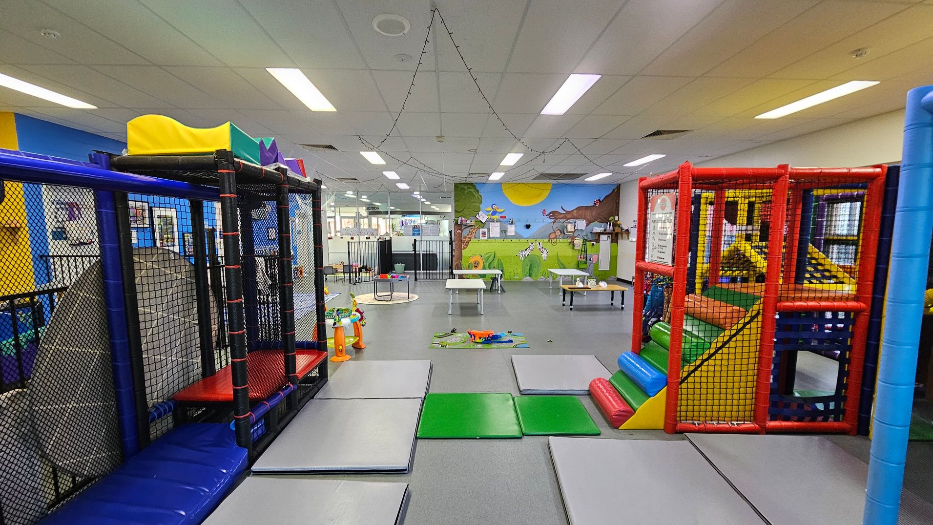 A colourful indoor play area with padded climbing structures on the left and right, featuring netted enclosures and slides. The floor is covered with grey and green mats. In the middle, there are small tables and toys scattered around. The back wall has a vibrant mural depicting a sunny outdoor scene with animals. The ceiling is lined with fluorescent lights and string lights. A colourful indoor play area with padded climbing structures on the left and right, featuring netted enclosures and slides. The floor is covered with grey and green mats. In the middle, there are small tables and toys scattered around. The back wall has a vibrant mural depicting a sunny outdoor scene with animals. The ceiling is lined with fluorescent lights and string lights.