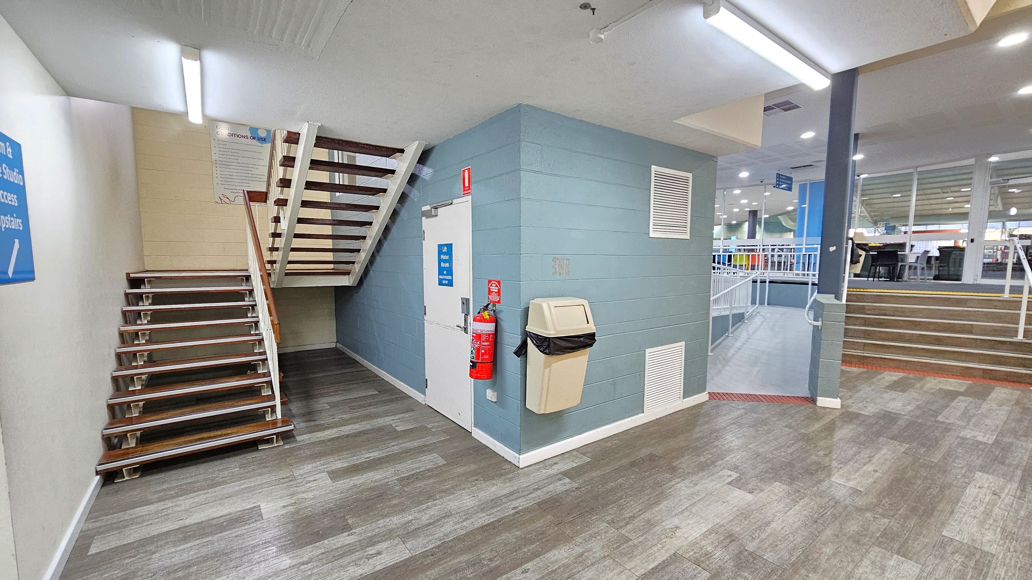 A hallway with a staircase on the left leading upwards, featuring wooden steps and white railings. In the centre, a blue wall with a white door, a fire extinguisher, and a beige bin attached. The floor is covered with grey and green patterned tiles. On the right, another set of stairs with a metal railing leads to an open area with tables and chairs. The ceiling has fluorescent lights. A hallway with a staircase on the left leading upwards, featuring wooden steps and white railings. In the centre, a blue wall with a white door, a fire extinguisher, and a beige bin attached. The floor is covered with grey and green patterned tiles. On the right, another set of stairs with a metal railing leads to an open area with tables and chairs. The ceiling has fluorescent lights.