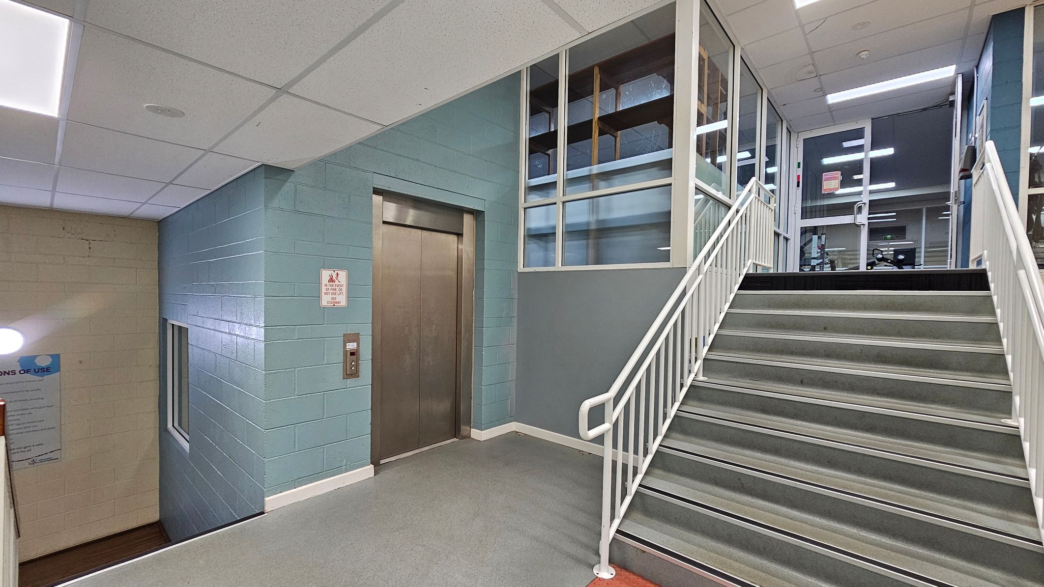 A stairwell with grey steps and white railings leads upwards to a glass door. To the left, there is a metal lift door set in a light blue wall. Above the lift, there are windows with a view into another room. The ceiling is made of white tiles with fluorescent lighting. The floor is grey, and there is a sign next to the lift. A small window is visible on the left wall, which is beige. A stairwell with grey steps and white railings leads upwards to a glass door. To the left, there is a metal lift door set in a light blue wall. Above the lift, there are windows with a view into another room. The ceiling is made of white tiles with fluorescent lighting. The floor is grey, and there is a sign next to the lift. A small window is visible on the left wall, which is beige.