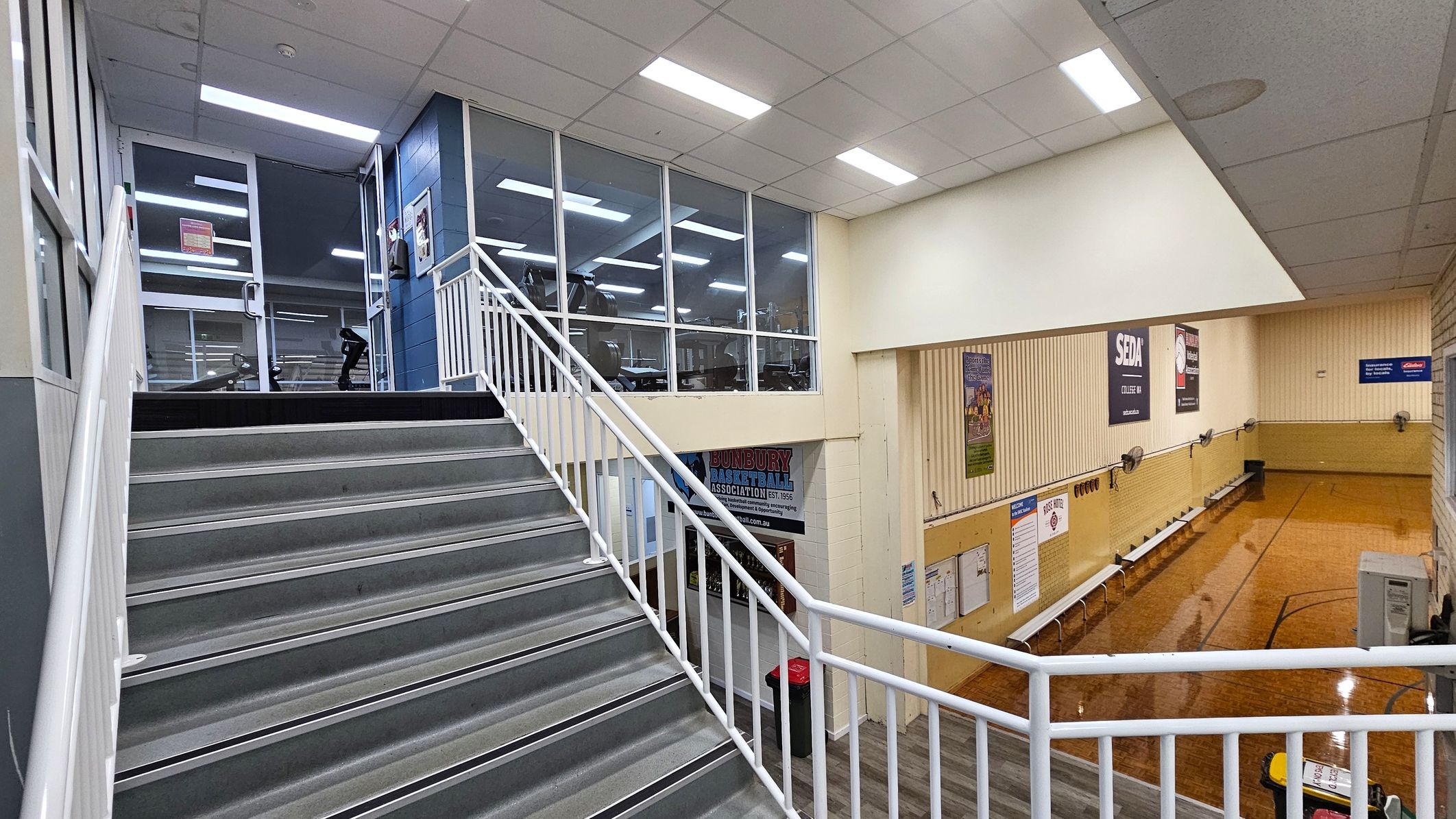 A staircase with grey steps and white railings leads up to a glass-walled area on the left, where gym equipment is visible. On the right, a basketball court with wooden flooring is partially flooded. The walls are adorned with various posters and signs. The ceiling is fitted with fluorescent lights, and the floor below the staircase is wooden. A staircase with grey steps and white railings leads up to a glass-walled area on the left, where gym equipment is visible. On the right, a basketball court with wooden flooring is partially flooded. The walls are adorned with various posters and signs. The ceiling is fitted with fluorescent lights, and the floor below the staircase is wooden.