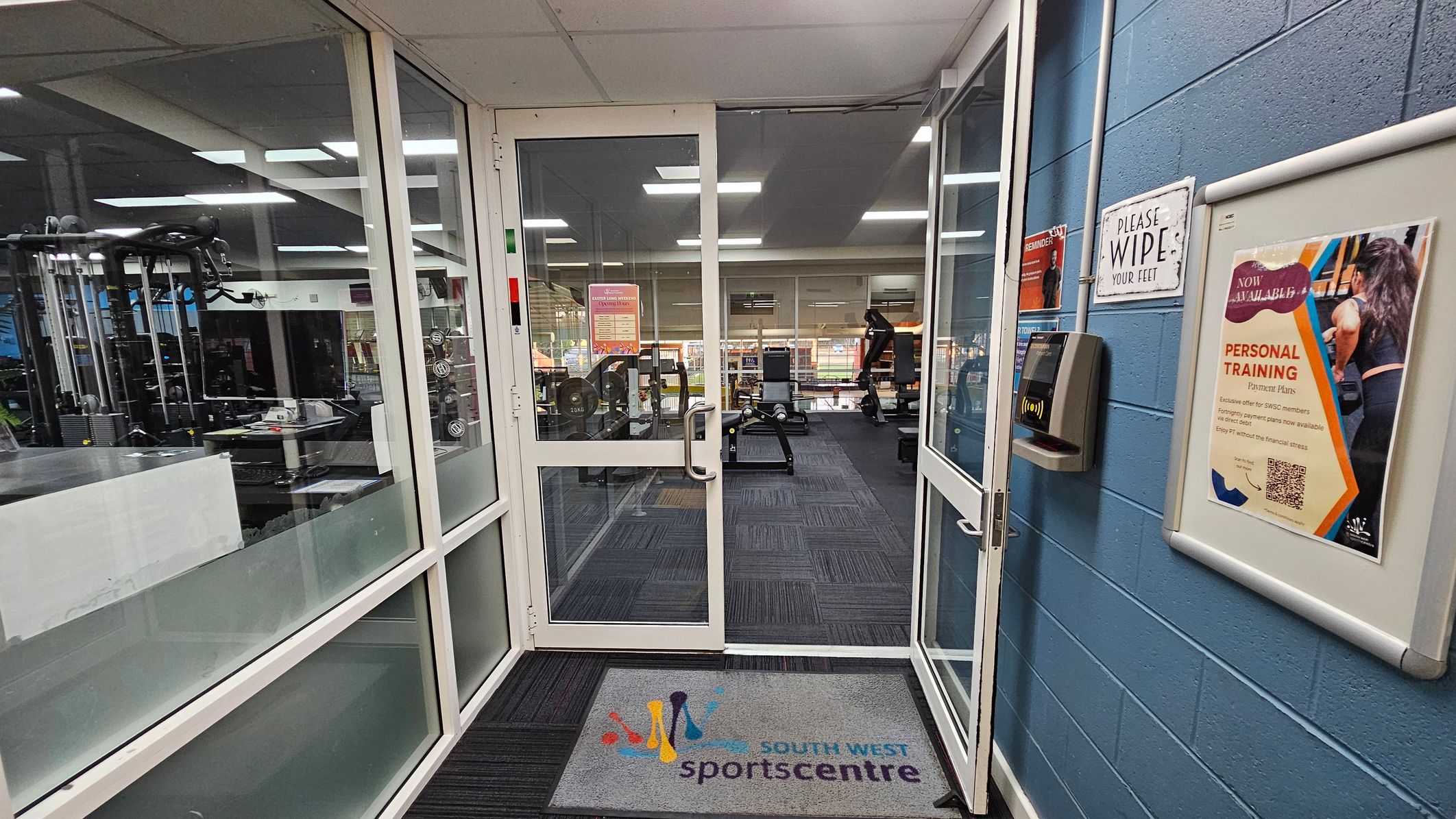 A gym entrance with glass doors and a mat on the floor labelled "South West Sports Centre". On the left, gym equipment is visible through large windows. On the right, a blue wall features a sign saying "PLEASE WIPE YOUR FEET" and a poster advertising personal training. A card reader is mounted on the wall. Inside, the gym has various exercise machines and equipment, with a carpeted floor and a ceiling with fluorescent lights. A gym entrance with glass doors and a mat on the floor labelled "South West Sports Centre". On the left, gym equipment is visible through large windows. On the right, a blue wall features a sign saying "PLEASE WIPE YOUR FEET" and a poster advertising personal training. A card reader is mounted on the wall. Inside, the gym has various exercise machines and equipment, with a carpeted floor and a ceiling with fluorescent lights.