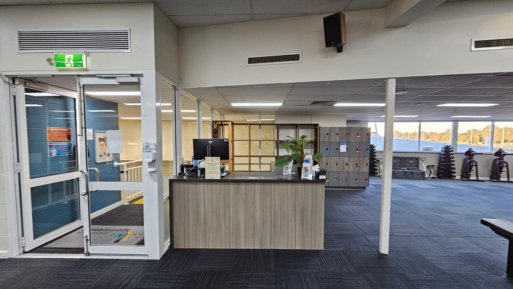 A reception area with a wooden desk in the centre, featuring a computer monitor, brochures, and a potted plant. To the left, a glass door with an illuminated exit sign above it leads to a hallway. On the right, a row of lockers is visible, along with exercise equipment like stationary bikes and stacked weights. The floor is carpeted in dark and light blue squares, and the ceiling has fluorescent lighting. Large windows in the background let in natural light. A reception area with a wooden desk in the centre, featuring a computer monitor, brochures, and a potted plant. To the left, a glass door with an illuminated exit sign above it leads to a hallway. On the right, a row of lockers is visible, along with exercise equipment like stationary bikes and stacked weights. The floor is carpeted in dark and light blue squares, and the ceiling has fluorescent lighting. Large windows in the background let in natural light.