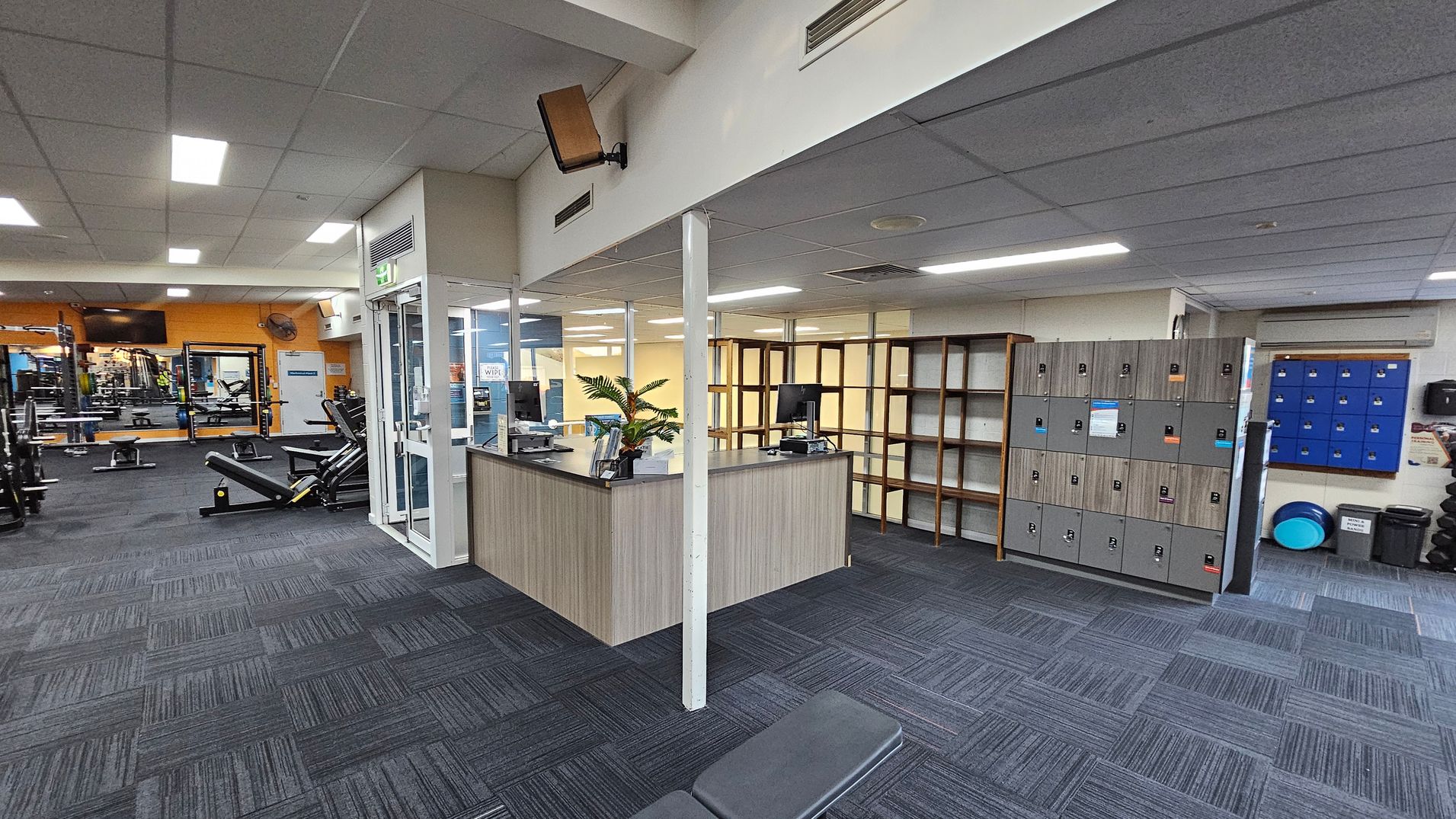 A gym interior with a reception desk in the centre, featuring a computer and a plant. To the right, there are grey lockers and blue lockers on the wall. The left side has gym equipment, including benches and weight racks. The floor is covered with patterned carpet tiles in shades of grey and purple. The ceiling is fitted with fluorescent lights. A gym interior with a reception desk in the centre, featuring a computer and a plant. To the right, there are grey lockers and blue lockers on the wall. The left side has gym equipment, including benches and weight racks. The floor is covered with patterned carpet tiles in shades of grey and purple. The ceiling is fitted with fluorescent lights.