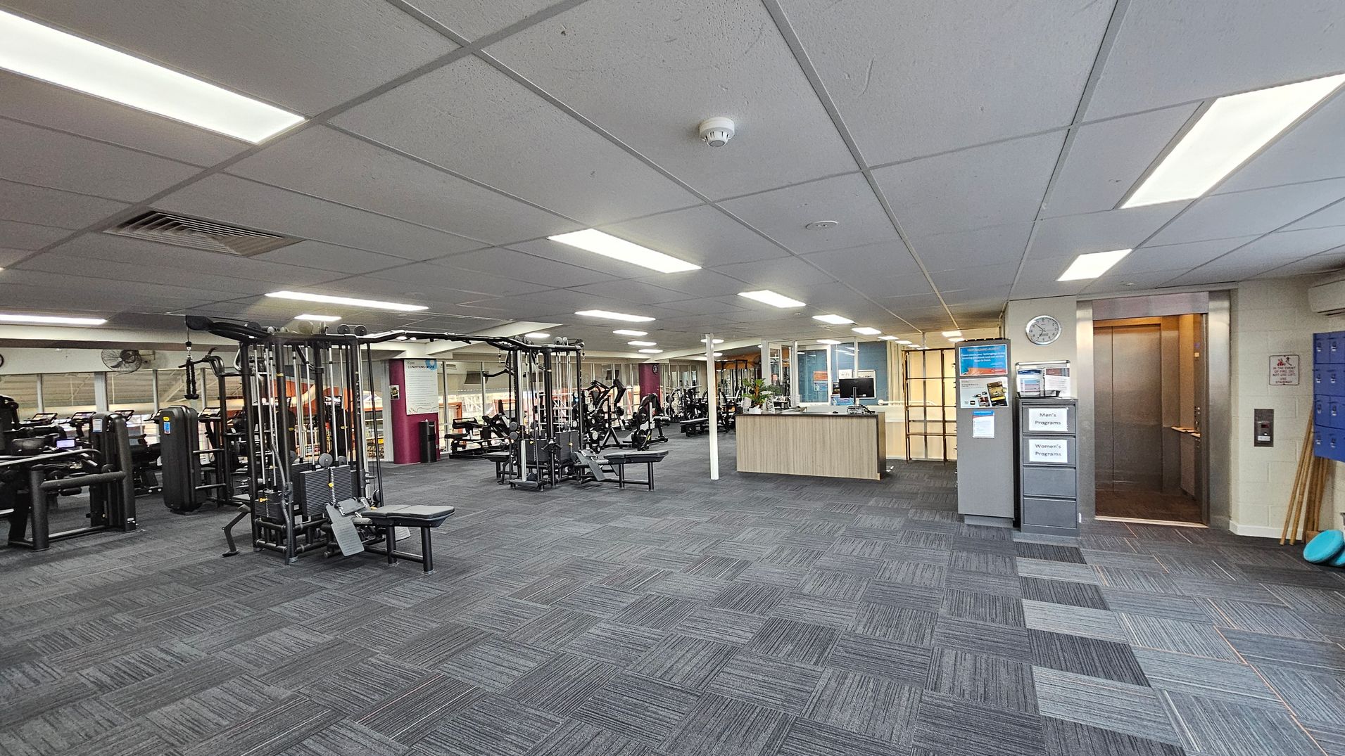 A spacious gym interior with a grey carpeted floor and a white panelled ceiling with fluorescent lights. On the left, there are various exercise machines, including weightlifting equipment. In the centre, a wooden reception desk is visible. On the right, there is an open lift with a clock above it and a set of blue lockers nearby. The walls are light-coloured, and there are informational signs and posters displayed. A spacious gym interior with a grey carpeted floor and a white panelled ceiling with fluorescent lights. On the left, there are various exercise machines, including weightlifting equipment. In the centre, a wooden reception desk is visible. On the right, there is an open lift with a clock above it and a set of blue lockers nearby. The walls are light-coloured, and there are informational signs and posters displayed.