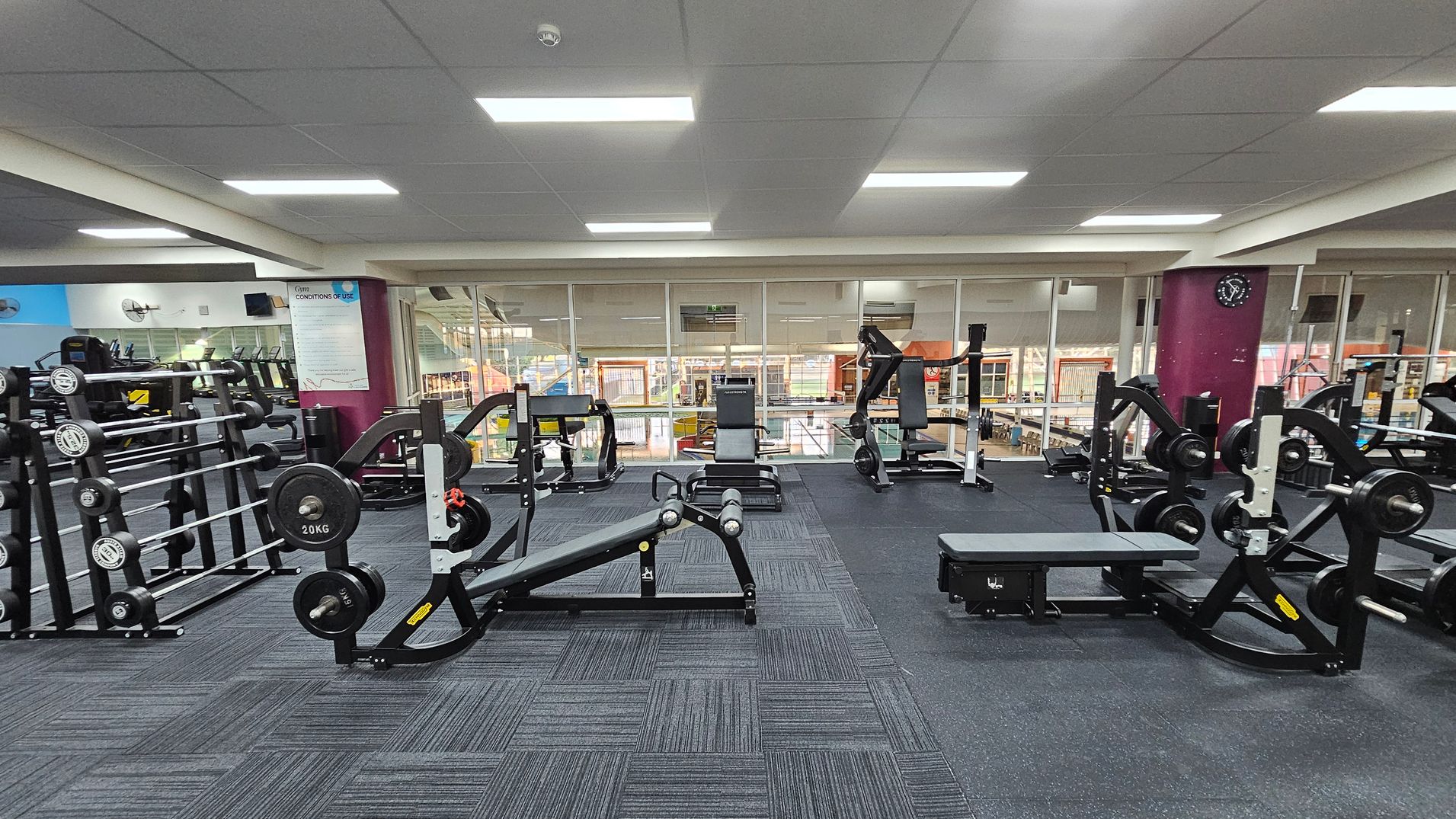 A gym with various weightlifting equipment. On the left, there are racks of dumbbells and barbells. In the centre, there are benches and weight machines. On the right, more weight machines are visible. The floor is covered with dark, patterned carpet tiles. The ceiling is fitted with fluorescent lights. The background features large windows overlooking another area of the gym, and a wall clock is mounted on a purple column. A gym with various weightlifting equipment. On the left, there are racks of dumbbells and barbells. In the centre, there are benches and weight machines. On the right, more weight machines are visible. The floor is covered with dark, patterned carpet tiles. The ceiling is fitted with fluorescent lights. The background features large windows overlooking another area of the gym, and a wall clock is mounted on a purple column.