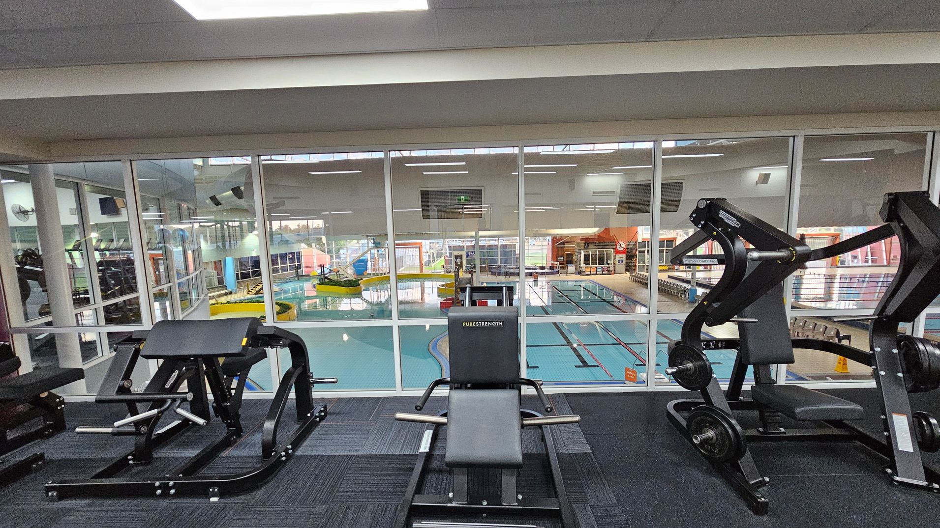 In the foreground, there are various gym machines on a dark carpeted floor. The middle features a large glass wall separating the gym area from an indoor swimming pool. The pool area has lanes and a yellow water slide. The ceiling is white with fluorescent lights. The background shows more gym equipment and a colourful pool area with windows allowing natural light. In the foreground, there are various gym machines on a dark carpeted floor. The middle features a large glass wall separating the gym area from an indoor swimming pool. The pool area has lanes and a yellow water slide. The ceiling is white with fluorescent lights. The background shows more gym equipment and a colourful pool area with windows allowing natural light.