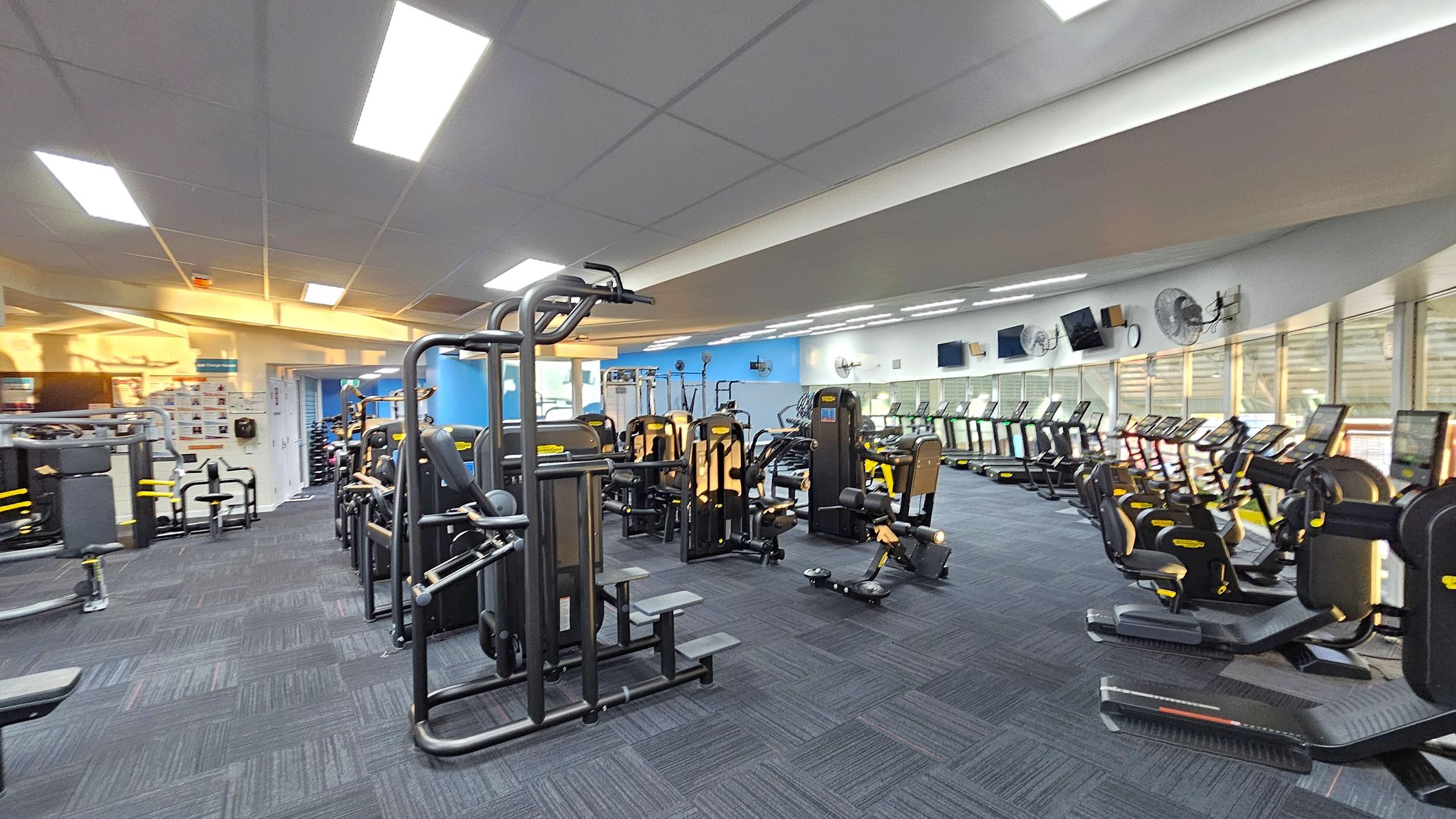 A spacious gym interior with a variety of exercise equipment. On the left, there are weight machines and benches. In the centre, more weight machines are arranged in rows. On the right, a line of treadmills and exercise bikes faces large windows. The floor is covered with dark blue carpet tiles, and the ceiling has bright fluorescent lights. The walls are painted blue and white, with several fans and televisions mounted above the treadmills. A spacious gym interior with a variety of exercise equipment. On the left, there are weight machines and benches. In the centre, more weight machines are arranged in rows. On the right, a line of treadmills and exercise bikes faces large windows. The floor is covered with dark blue carpet tiles, and the ceiling has bright fluorescent lights. The walls are painted blue and white, with several fans and televisions mounted above the treadmills.