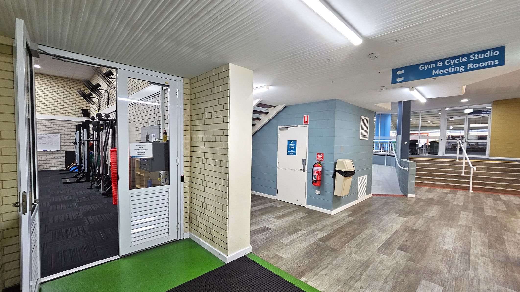 A gym entrance is on the left with glass doors and visible exercise equipment inside. The floor is green near the entrance and transitions to a wood-like pattern in the hallway. A white door with a blue sign is in the centre, next to a fire extinguisher and a bin. Stairs with a white railing lead up on the right. The ceiling is white with fluorescent lights. A blue sign above points to the gym, cycle studio, and meeting rooms. A gym entrance is on the left with glass doors and visible exercise equipment inside. The floor is green near the entrance and transitions to a wood-like pattern in the hallway. A white door with a blue sign is in the centre, next to a fire extinguisher and a bin. Stairs with a white railing lead up on the right. The ceiling is white with fluorescent lights. A blue sign above points to the gym, cycle studio, and meeting rooms.