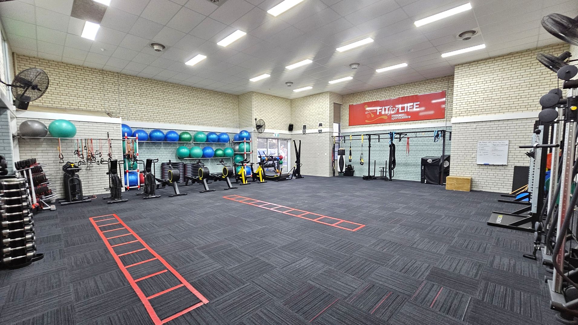 A spacious gym with a dark carpeted floor featuring red agility ladders. On the left, there are stacks of dumbbells, exercise bikes, and a variety of exercise balls on shelves. The right side has more equipment, including resistance bands and a whiteboard. The back wall displays a large red "Fit for Life" banner. The ceiling is fitted with bright fluorescent lights, and the walls are made of light-coloured bricks. A spacious gym with a dark carpeted floor featuring red agility ladders. On the left, there are stacks of dumbbells, exercise bikes, and a variety of exercise balls on shelves. The right side has more equipment, including resistance bands and a whiteboard. The back wall displays a large red "Fit for Life" banner. The ceiling is fitted with bright fluorescent lights, and the walls are made of light-coloured bricks.