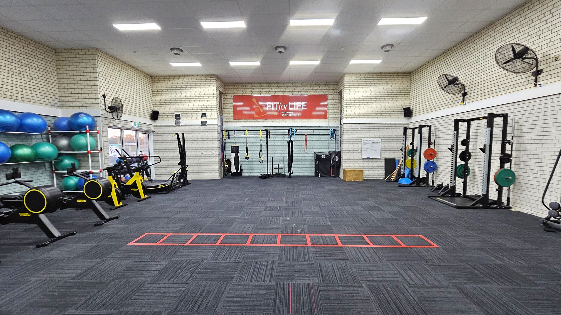 A gym with a dark carpeted floor and white brick walls. On the left, there are shelves with blue and green exercise balls and two rowing machines with yellow accents. In the centre, a red sign reads "FIT for LIFE" above various hanging exercise equipment. On the right, there are weightlifting racks with colourful weight plates. Ceiling fans are mounted on the walls, and the ceiling has bright rectangular lights. A gym with a dark carpeted floor and white brick walls. On the left, there are shelves with blue and green exercise balls and two rowing machines with yellow accents. In the centre, a red sign reads "FIT for LIFE" above various hanging exercise equipment. On the right, there are weightlifting racks with colourful weight plates. Ceiling fans are mounted on the walls, and the ceiling has bright rectangular lights.