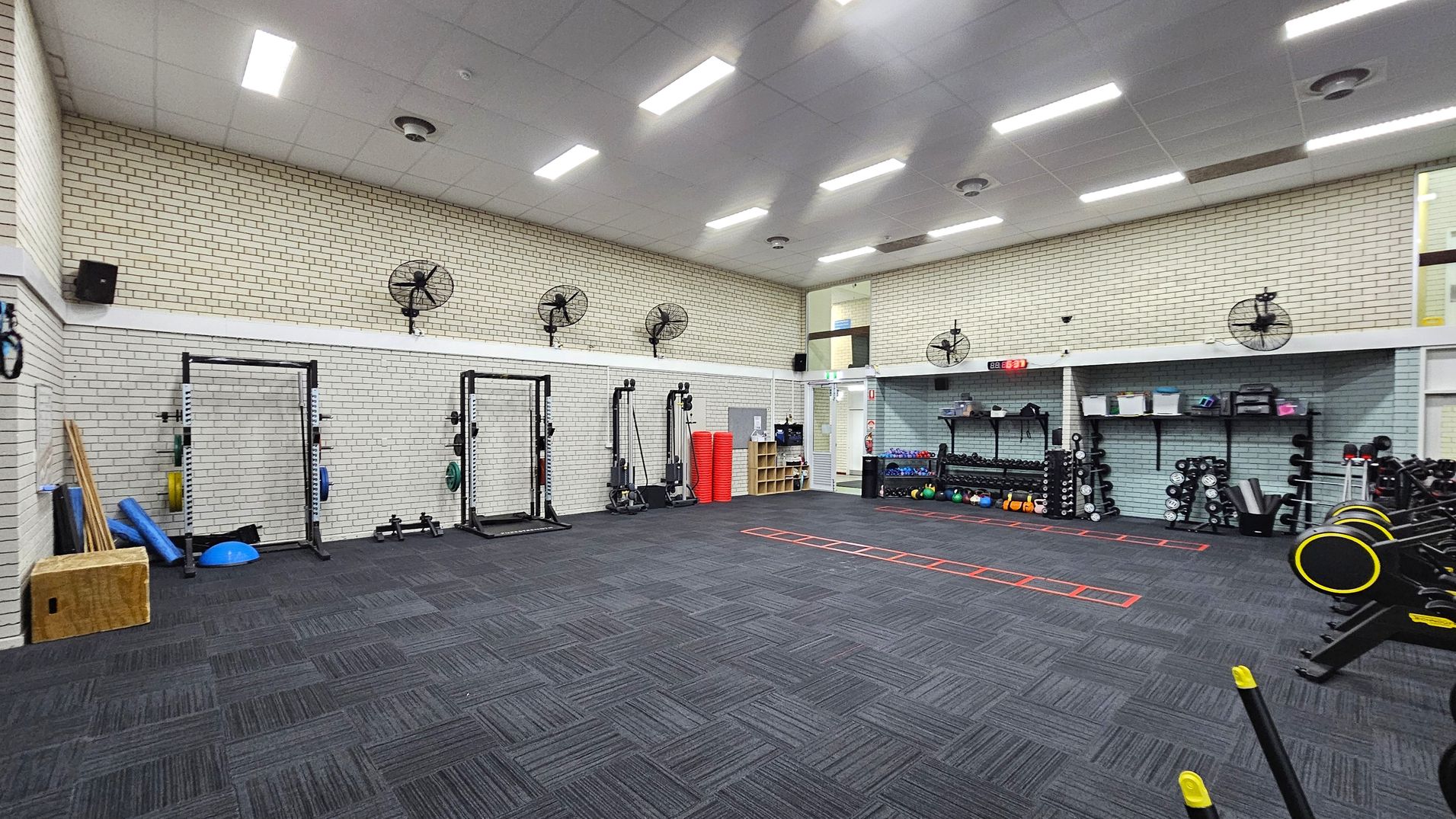 A spacious gym with a dark carpeted floor featuring a mix of grey and purple tones. The left side has exercise equipment like squat racks, weights, and foam rollers. The right side has shelves with kettlebells, dumbbells, and other fitness gear. The walls are light-coloured brick with several wall-mounted fans. The ceiling is high with bright fluorescent lights. A red agility ladder is laid out on the floor. A spacious gym with a dark carpeted floor featuring a mix of grey and purple tones. The left side has exercise equipment like squat racks, weights, and foam rollers. The right side has shelves with kettlebells, dumbbells, and other fitness gear. The walls are light-coloured brick with several wall-mounted fans. The ceiling is high with bright fluorescent lights. A red agility ladder is laid out on the floor.