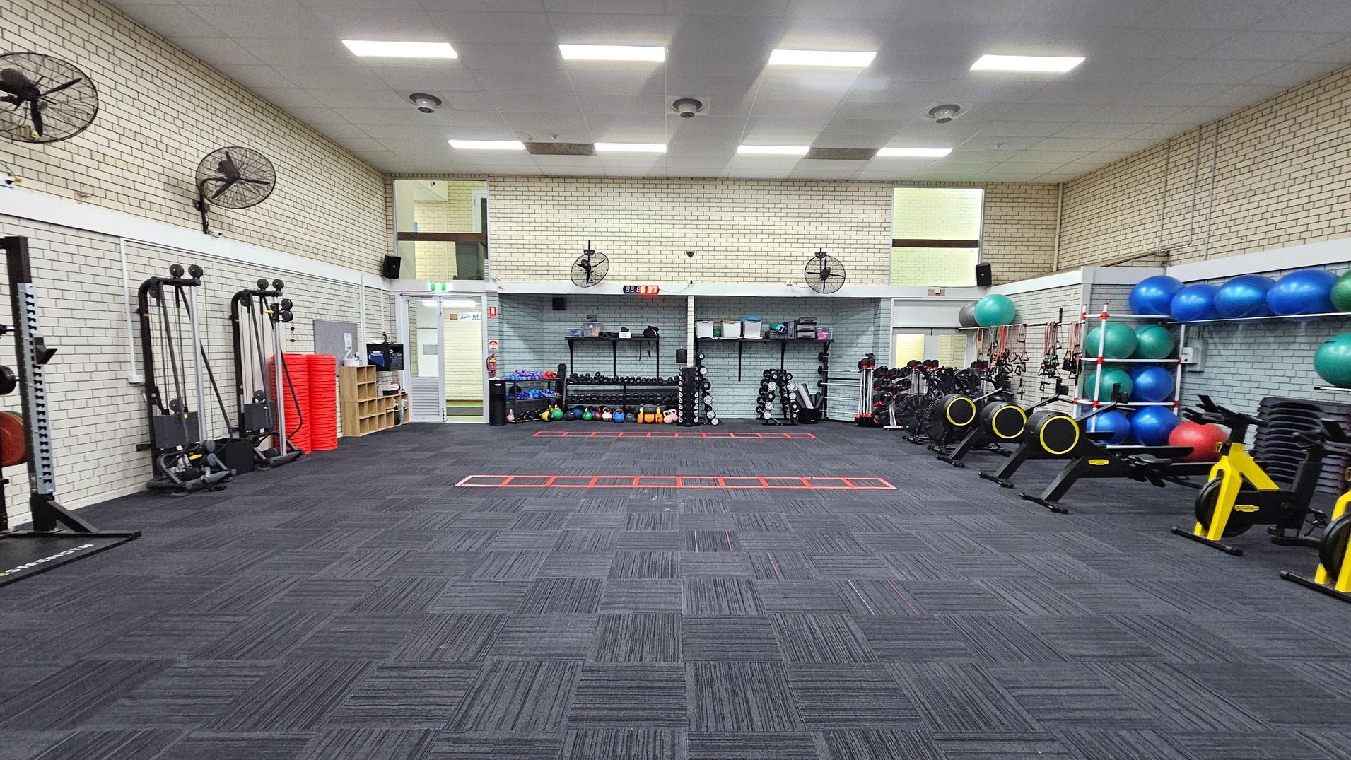 A spacious gym with a dark carpeted floor and a white tiled ceiling. On the left, there are exercise machines and red mats stacked against the wall. The back wall features shelves with kettlebells, dumbbells, and other equipment. On the right, there are exercise bikes and shelves with blue and green exercise balls. Wall-mounted fans are positioned around the room. The walls are light-coloured brick, and there are fluorescent lights on the ceiling. A spacious gym with a dark carpeted floor and a white tiled ceiling. On the left, there are exercise machines and red mats stacked against the wall. The back wall features shelves with kettlebells, dumbbells, and other equipment. On the right, there are exercise bikes and shelves with blue and green exercise balls. Wall-mounted fans are positioned around the room. The walls are light-coloured brick, and there are fluorescent lights on the ceiling.