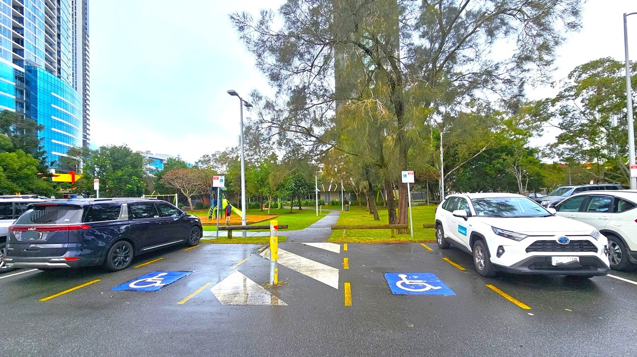 A parking area with two cars parked in designated disabled spaces, marked by blue signs on the ground. On the left, a dark-coloured vehicle, and on the right, a white SUV. In the background, a park with trees, a pathway, and a playground. A tall building with blue glass windows is visible on the left. The ground is wet, suggesting recent rain, and there are street lamps and signs around the area.