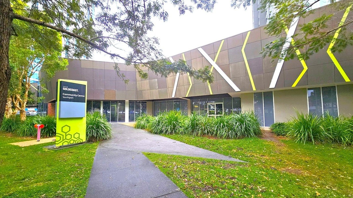 A modern building with a geometric facade featuring grey panels and diagonal white and yellow accents. In the foreground, a path leads to the entrance, flanked by lush green grass and plants. On the left, a sign reads "City of Gold Coast Community Centre Southport" in white text on a blue and green background. Trees with overhanging branches frame the scene, adding a natural element to the urban setting.