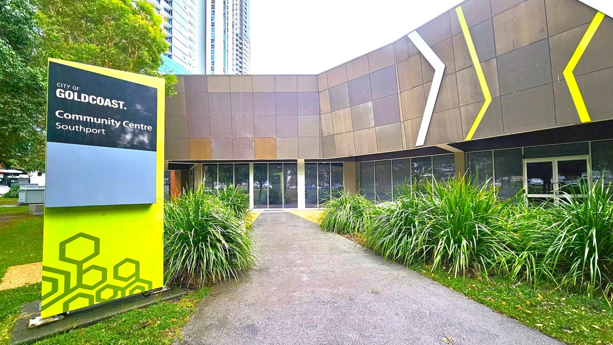 A modern building with a geometric facade featuring grey and brown panels, accented by white and yellow diagonal lines. In the foreground, a path flanked by lush green plants leads to the entrance. On the left, a large sign reads "City of Gold Coast Community Centre Southport" with a green and blue design. Tall buildings are visible in the background, partially obscured by trees.