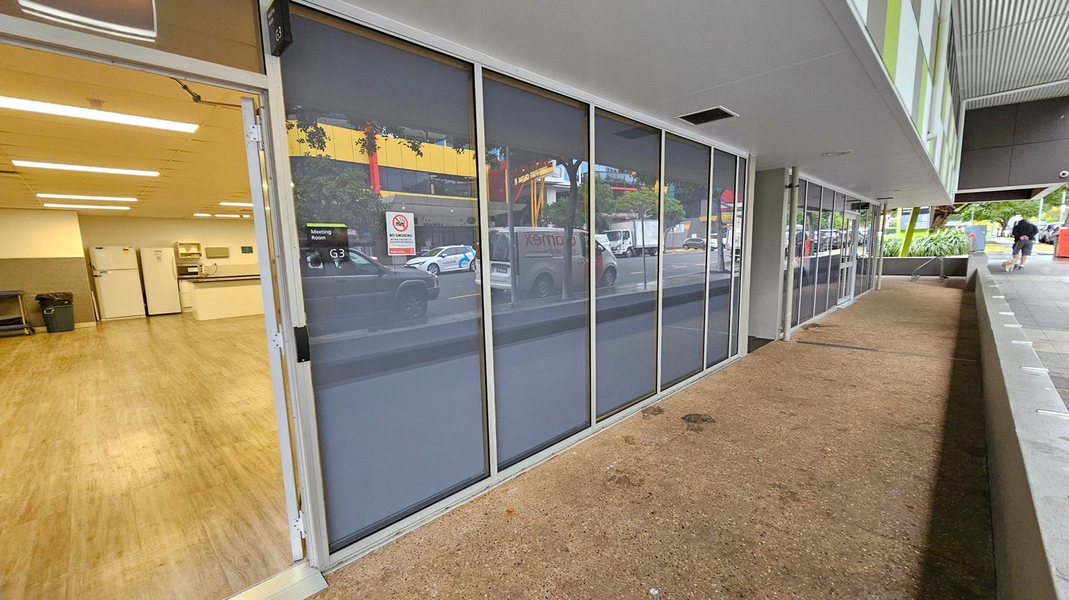 On the left, an open door reveals an interior space with wooden flooring, white appliances, and a counter. The right side features a row of large glass windows with reflections of vehicles and buildings outside. The ground is a textured concrete surface, and the ceiling is smooth and white. A sign on the door reads "Meeting Room G3". In the background, a person walks along a path bordered by greenery and a red post box.