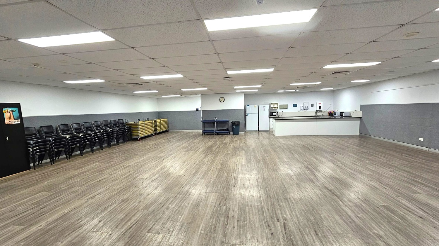 A spacious room with a wooden floor and a ceiling with fluorescent lights. On the left, there are stacks of black chairs and a black cabinet with a poster. In the middle, there are two trolleys and a clock on the wall. On the right, a kitchen area with a long counter, white appliances, including a fridge and microwave, and various kitchen items. The walls are white with a grey lower section.