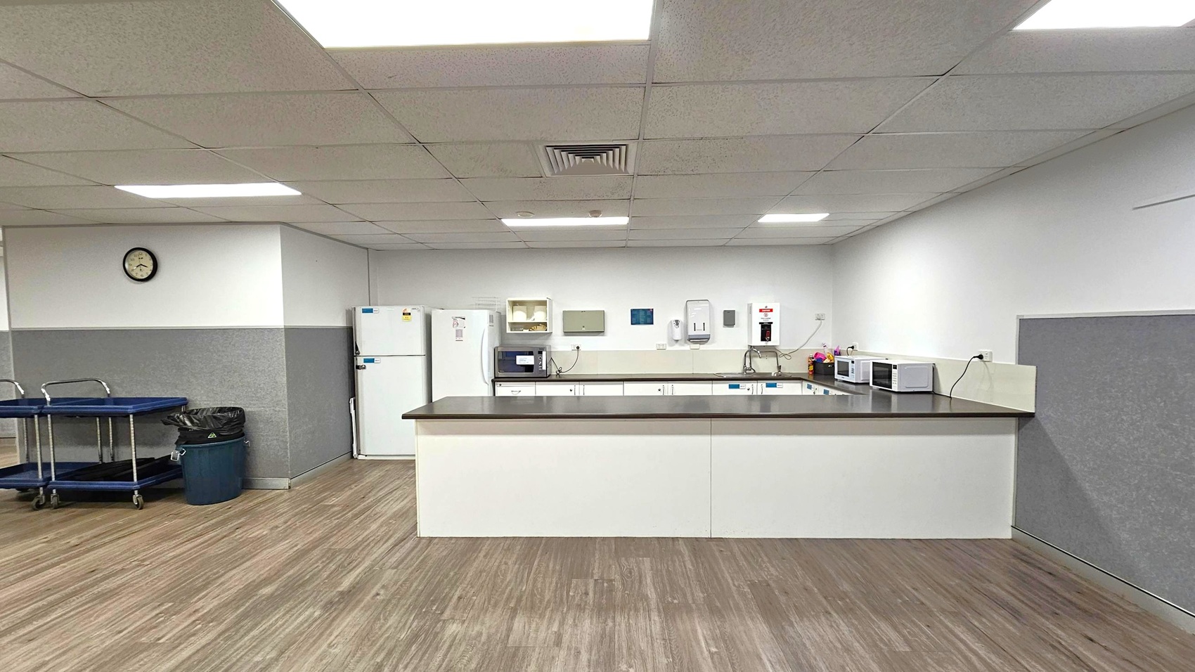 A kitchen area with a long counter in the centre, featuring multiple microwaves and a sink. On the left, there is a white refrigerator and a wall clock above it. A black rubbish bin and a trolley are also visible. The floor is wooden, and the ceiling has recessed lighting and a vent. The walls are half grey and half white, with various kitchen appliances and dispensers mounted on them.